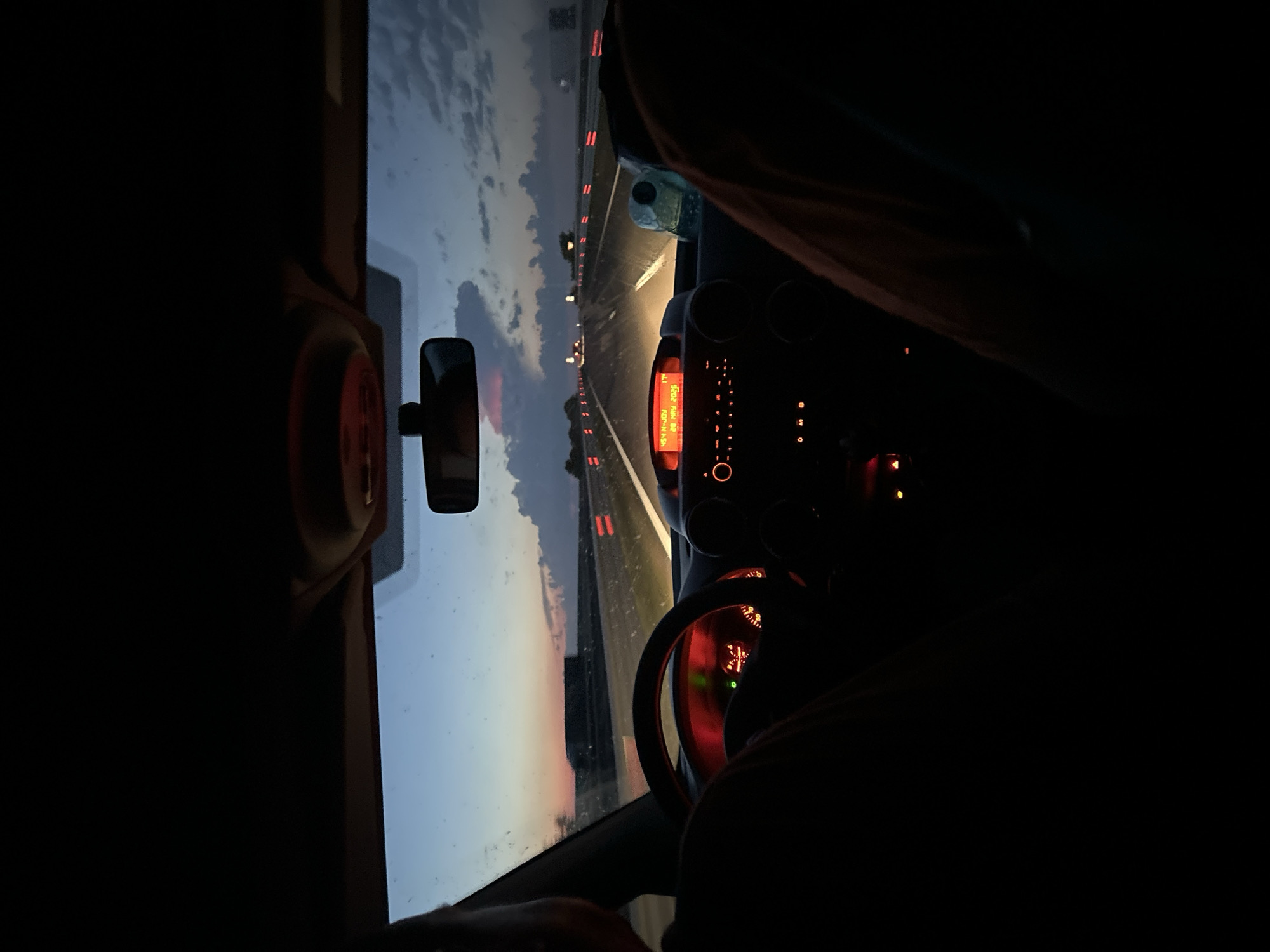 The image shows the interior of a car driving on a highway at dusk or dawn. The dashboard is visible, with the radio display lit up. The steering wheel and instrument panel are illuminated in red. Through the windshield, the highway stretches into the distance, marked with orange delineators. The sky is a mix of blue, gray, and pink hues. The location is not specified in the image context, only that the image should be related to Kozare, Bulgaria. No people are fully visible, only parts of what appears to be clothing belonging to the driver and a passenger.