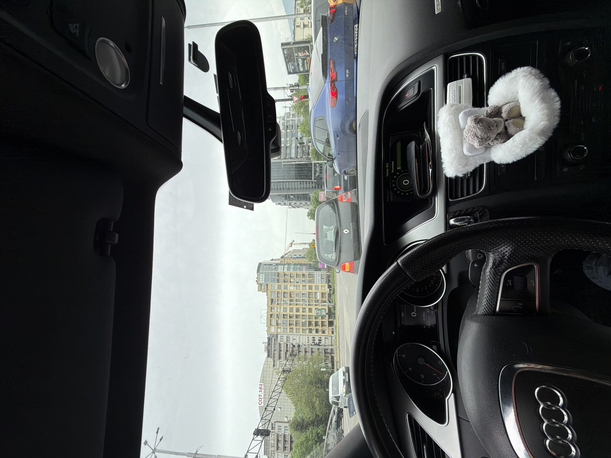 A view from the inside of an Audi car, likely an A5 model, stuck in traffic on a city street in Sofia, Bulgaria. The vehicle is facing an intersection with a traffic light visible. Outside, other cars, including a blue BMW X3 and a grey Renault Twingo, are also stopped in traffic. Tall buildings are visible in the background, along with a billboard advertising "BETWILD." A sign reads "COT 161." Inside the Audi, a heart-shaped, plush decoration with a small teddy bear is clipped to the air vent. The speedometer reads 0 km/h. The odometer reads 274222 and other information on the dashboard display is also visible, with text “Yettel BG” beside a number pad.
