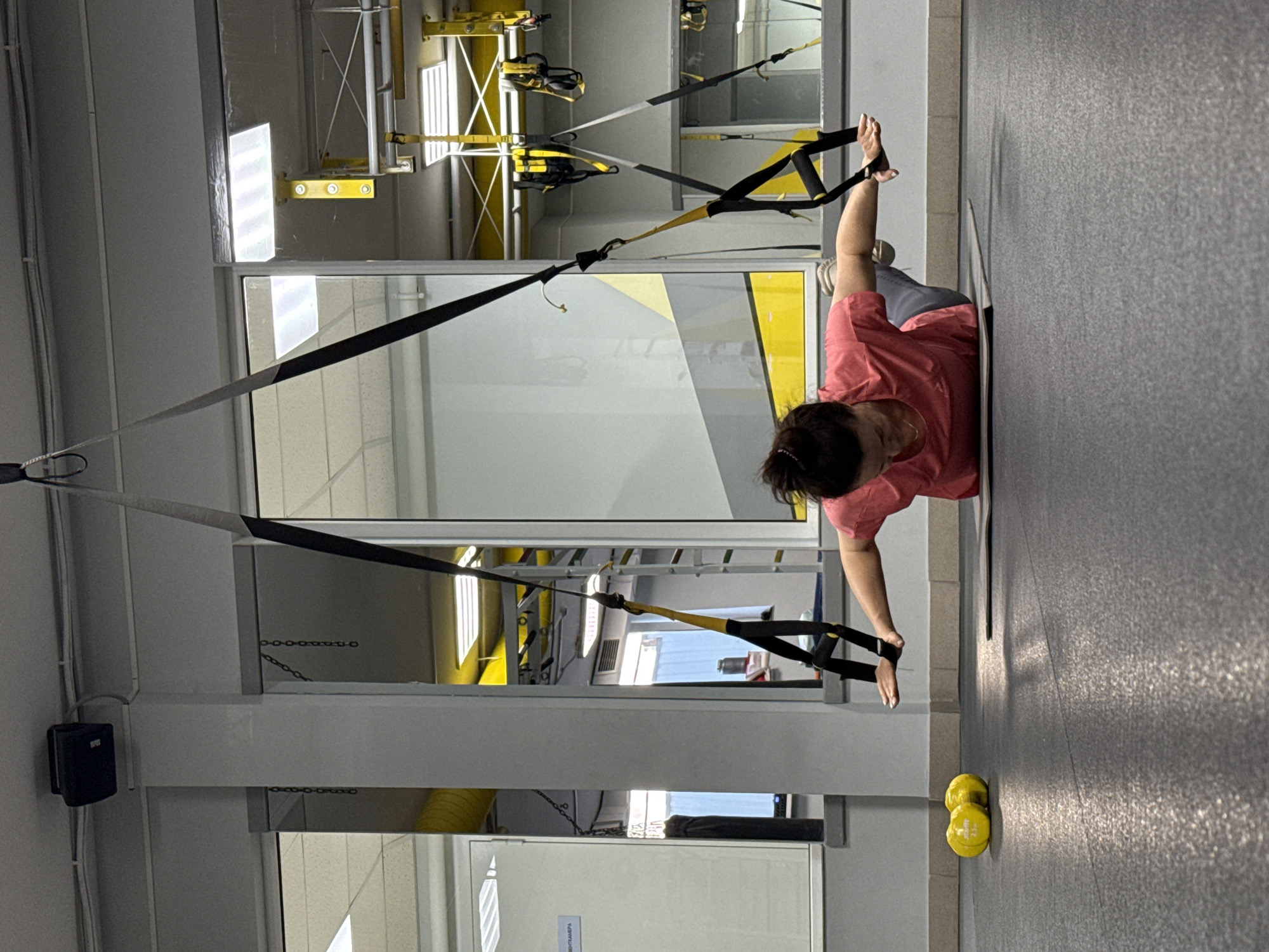 The image shows a woman doing a push-up exercise using TRX suspension straps in a gym. The gym has large windows, yellow accents, and fitness equipment. The location is the city of Chaikovsky, Russia. The woman is wearing a pink shirt and grey pants. A pair of yellow dumbbells are on the floor.