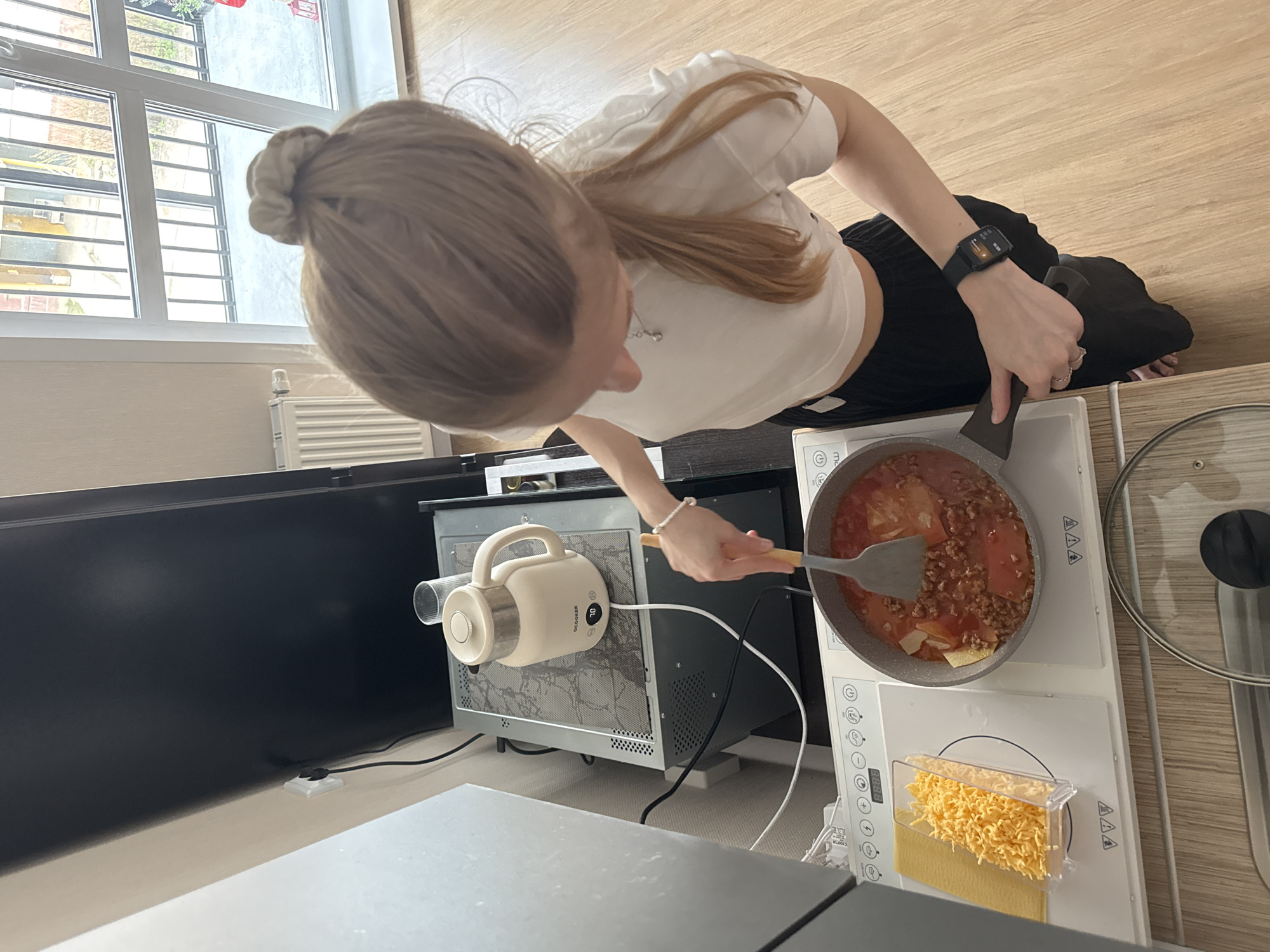An overhead shot captures a young woman with light brown hair, tied in a bun, focused on cooking in a brightly lit indoor setting, likely a kitchen or a compact living space. She wears a white cropped top and dark pants, with a black smartwatch on her left wrist displaying "10:55".
The woman is stirring a pan with a spatula, preparing a dish that appears to be lasagna or a similar pasta dish, containing ground meat, sauce, and flat pasta sheets. The pan sits on a white portable induction cooktop, which features various control buttons and symbols. Next to the pan, a clear container holds shredded cheese and uncooked lasagna sheets. A pot lid rests to the right of the cooktop.
Behind the cooking area, a beige "OCOOKER" brand electric kettle, displaying "70" on its digital screen, is placed on top of a dark-colored appliance, possibly a microwave or oven. Electrical cords are visible around these devices.
In the background, a large window with white frames and horizontal security bars allows ample daylight into the room. Outside the window, a glimpse of an adjacent building and some foliage can be seen. To the left, a large, dark screen, possibly a television or computer monitor, is visible. The floor is covered with light-colored wood laminate. The scene depicts a calm daytime cooking activity in what could be an apartment in Perm, Russia.