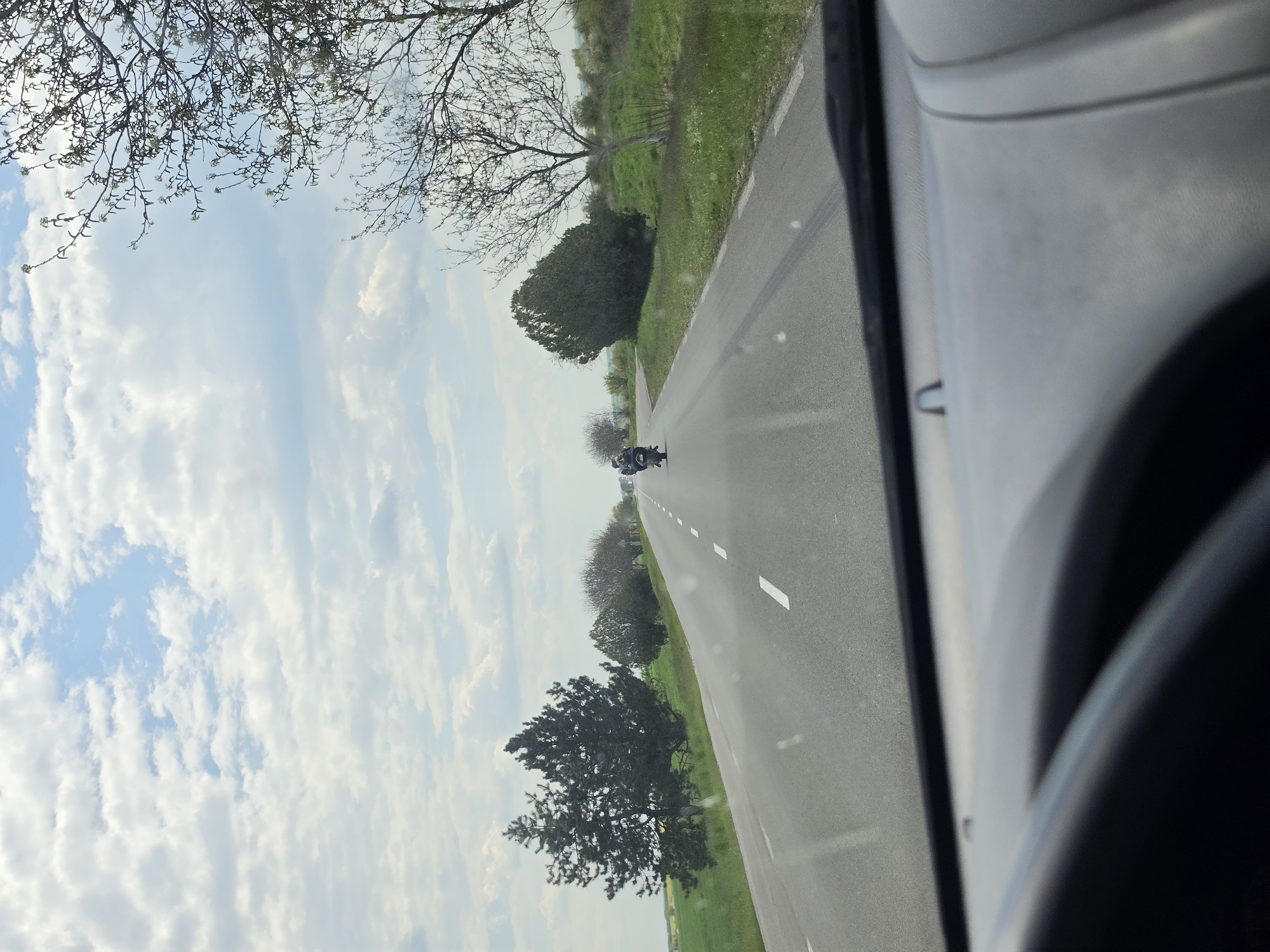 View from inside a vehicle traveling on a paved road outside Gorno Aleksandrovo, Bulgaria. The road has a white centerline and green fields on either side, lined with trees. A motorcycle is approaching in the distance. The sky is partly cloudy. The interior of the vehicle is visible on the right.