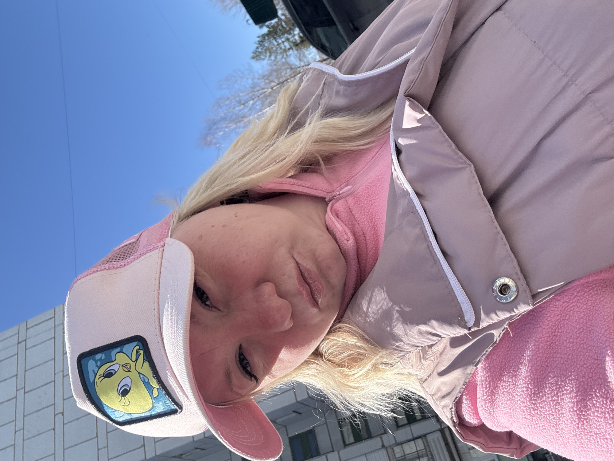 A medium shot shows a woman with blonde hair and blue eyes, wearing a pink Tweety Bird cap, a pink fleece jacket, and a light purple puffer jacket with white trim. She is facing the camera with a slight downward angle. In the background, there's a pale blue sky and parts of buildings. Based on geolocation, this image was taken in Chaikovsky, Perm Krai, Russia.