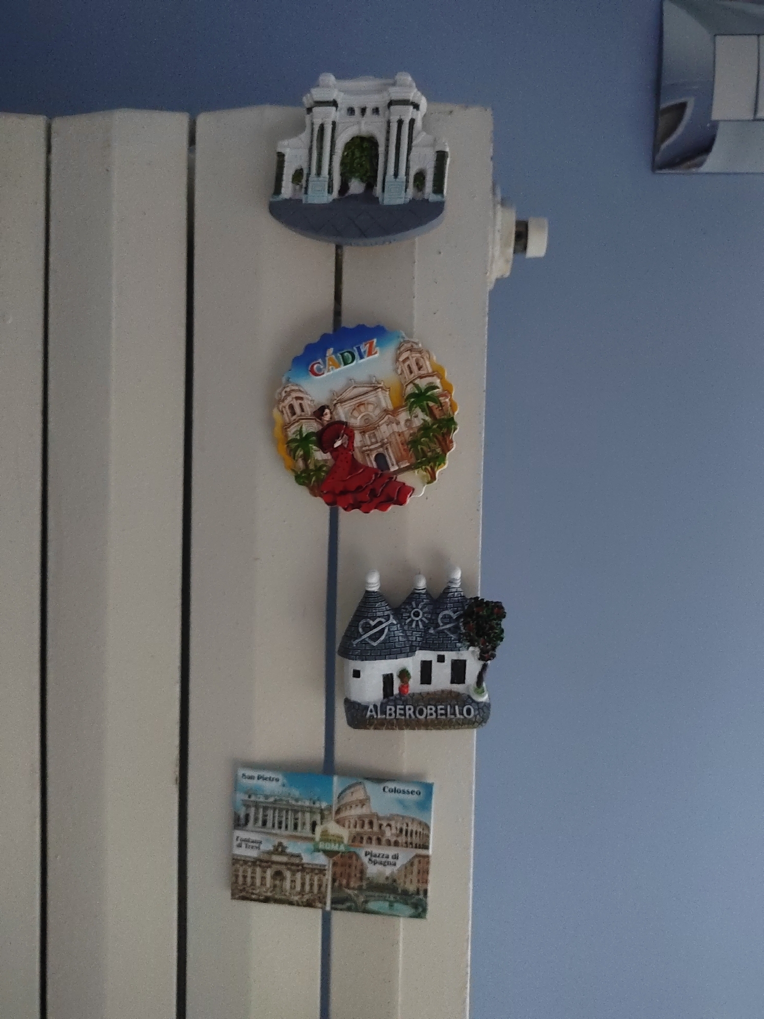 A vertical shot shows a radiator in an indoor setting, against a blue wall. Several refrigerator magnets are attached to the radiator. From top to bottom, the magnets depict: a white, architectural structure with an arched entrance and columns; a circular magnet with a flamenco dancer and the word "CADIZ"; a small white house with conical roofs, and the word "ALBEROBELLO" underneath; and a rectangular magnet showing two Italian landmarks, the Colosseum and what appears to be St. Peter's Basilica, with Italian text below. The radiator is white and has a ribbed texture. The lighting is even, and the image is in focus. No people or animals are present in the frame. The location is not Porto Recanati, Italy, but rather Cadiz, Spain and Alberobello and Rome, Italy based on the magnets.