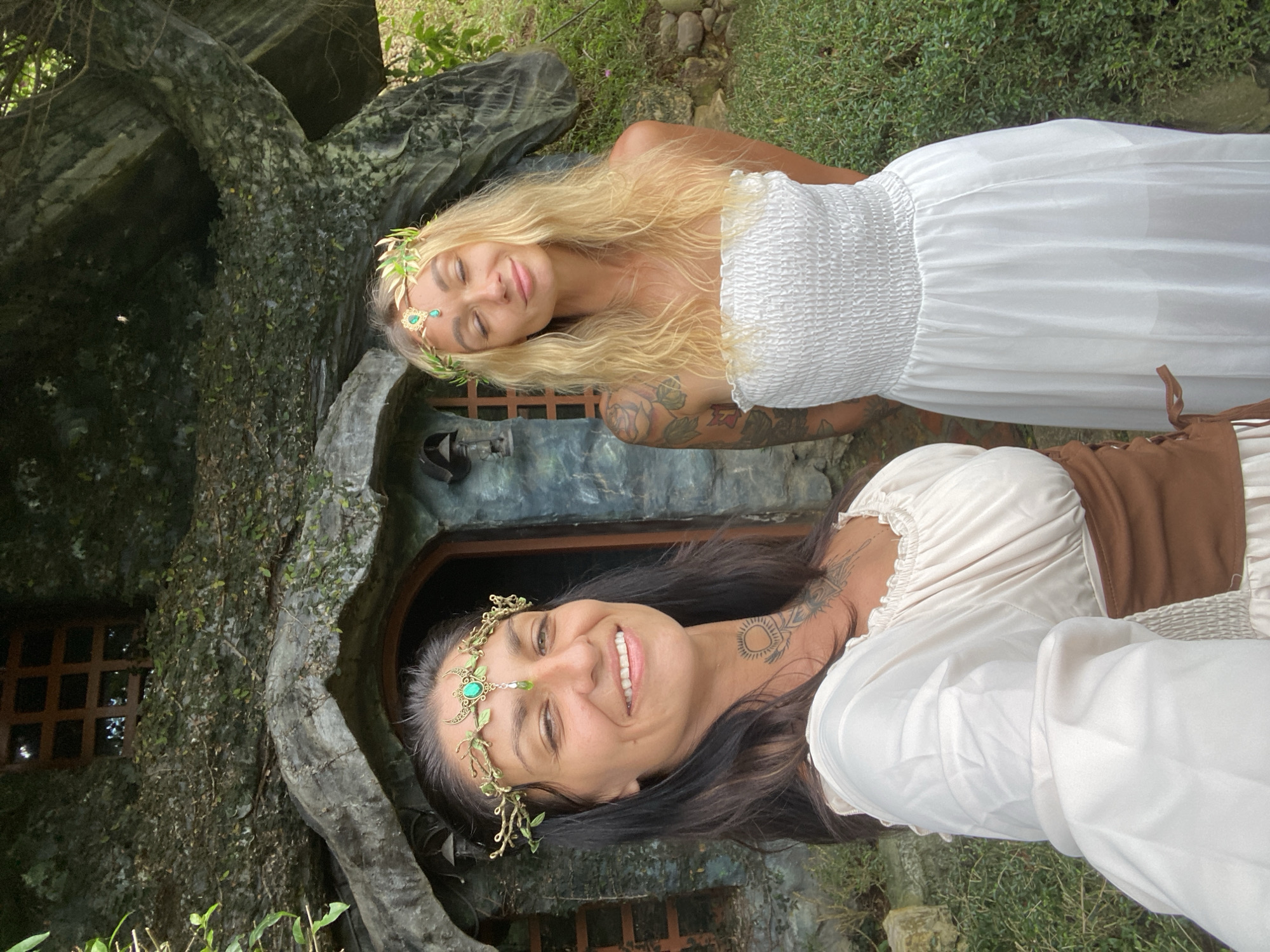 The image depicts two women dressed in ethereal, woodland-inspired attire, posing in front of a whimsical, tree-like structure that appears to be a dwelling. The woman on the left has dark hair and is wearing a cream-colored dress with a brown corset and a golden headdress adorned with green jewels and leaves. She has a tattoo on her neck. The woman on the right has blonde, wavy hair and is wearing a white strapless smocked dress and a similar golden headdress. She also has tattoos on her arms. The background features a structure with a dark wooden door and window, covered in moss and ivy, suggesting it is integrated with nature. The surrounding environment is lush and green, with dense foliage and a rustic stone path visible to the right. The lighting suggests it is daytime, possibly overcast, creating a soft, dreamlike atmosphere. The location is identified as Cam Ly - Da Lat, Vietnam.