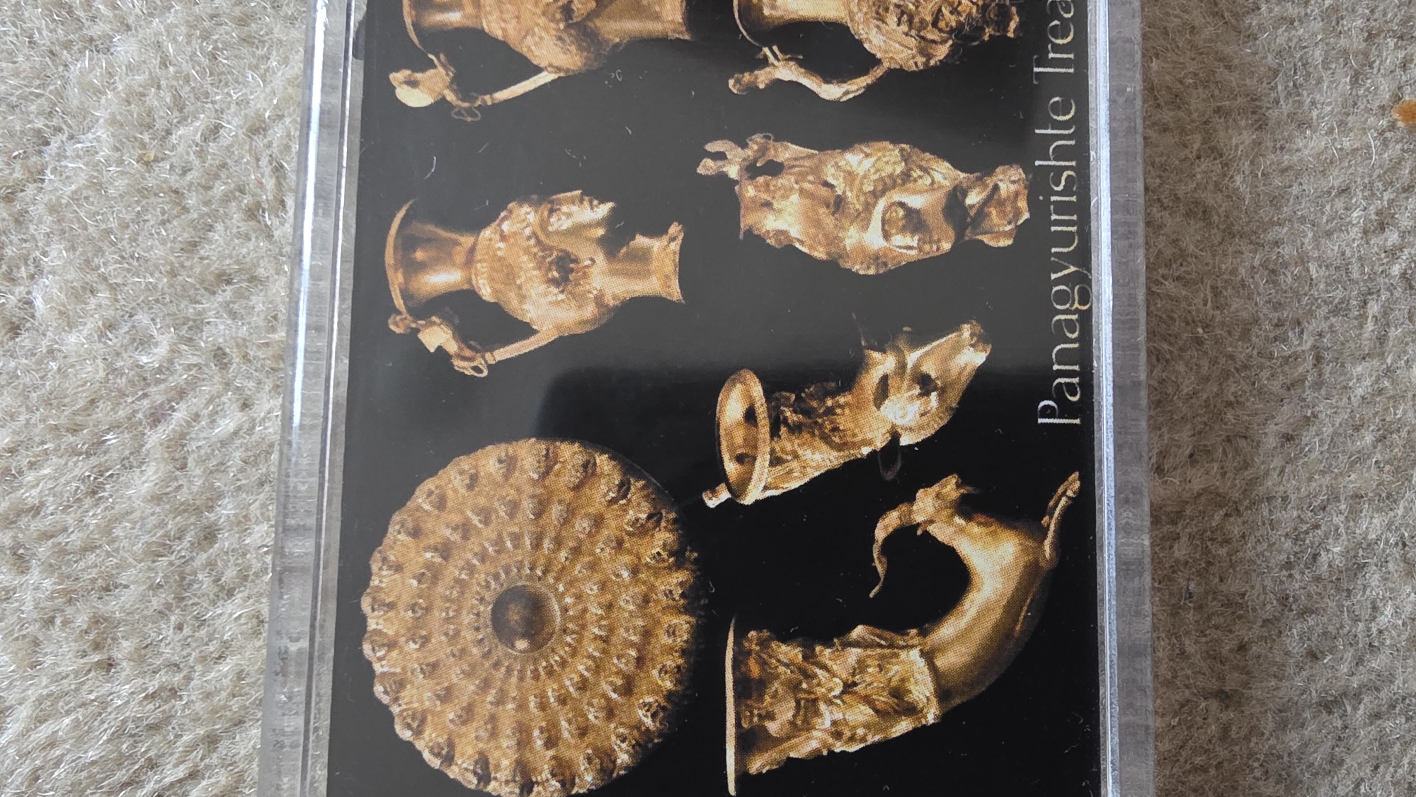 The image depicts a promotional item, potentially a postcard or souvenir, encased in a clear plastic sleeve. The background is a textured, neutral-colored surface, possibly carpet or fabric. The central focus of the image is the promotional item displaying a collection of golden artifacts against a black background. The artifacts appear to be of ancient origin, including vessels, animal-shaped rhytons (drinking horns), and a decorative plate. The text "Panagyurishte Trea" is partially visible on the right side of the image. Based on this text, the artifacts are likely part of the Panagyurishte Treasure, which is commonly associated with the area of Panagyurishte, Bulgaria. Blagoevgrad is another city in Bulgaria. While it's not directly associated with the Panagyurishte Treasure's origin, museums in Bulgaria, including those in Blagoevgrad, may exhibit replicas or information about the treasure.