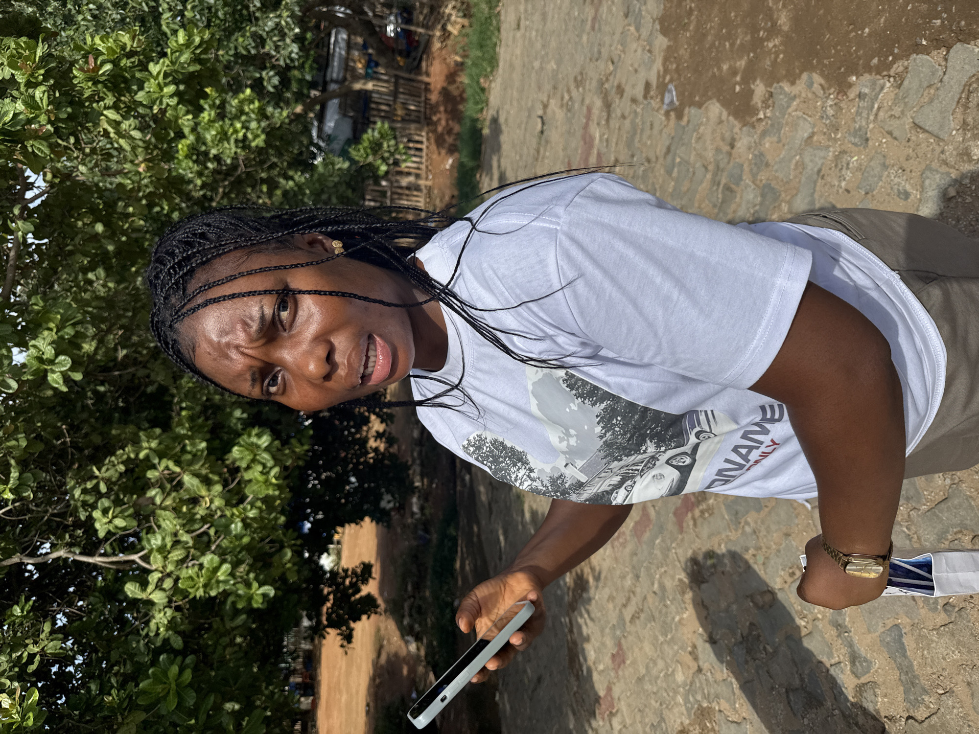 A woman with dark skin and black braided hair stands outdoors, looking to her right with a confused or concerned expression. She is wearing a white t-shirt with a graphic print and khaki pants. Her left hand holds a light-colored smartphone, and her right hand, adorned with a gold watch, clutches a white paper bag. The background features a large tree with lush green leaves, a cobblestone pathway, and a wooden fence. The scene appears to be a public outdoor space within Abuja, Nigeria, evidenced by the typical vegetation and paving commonly found in the city.