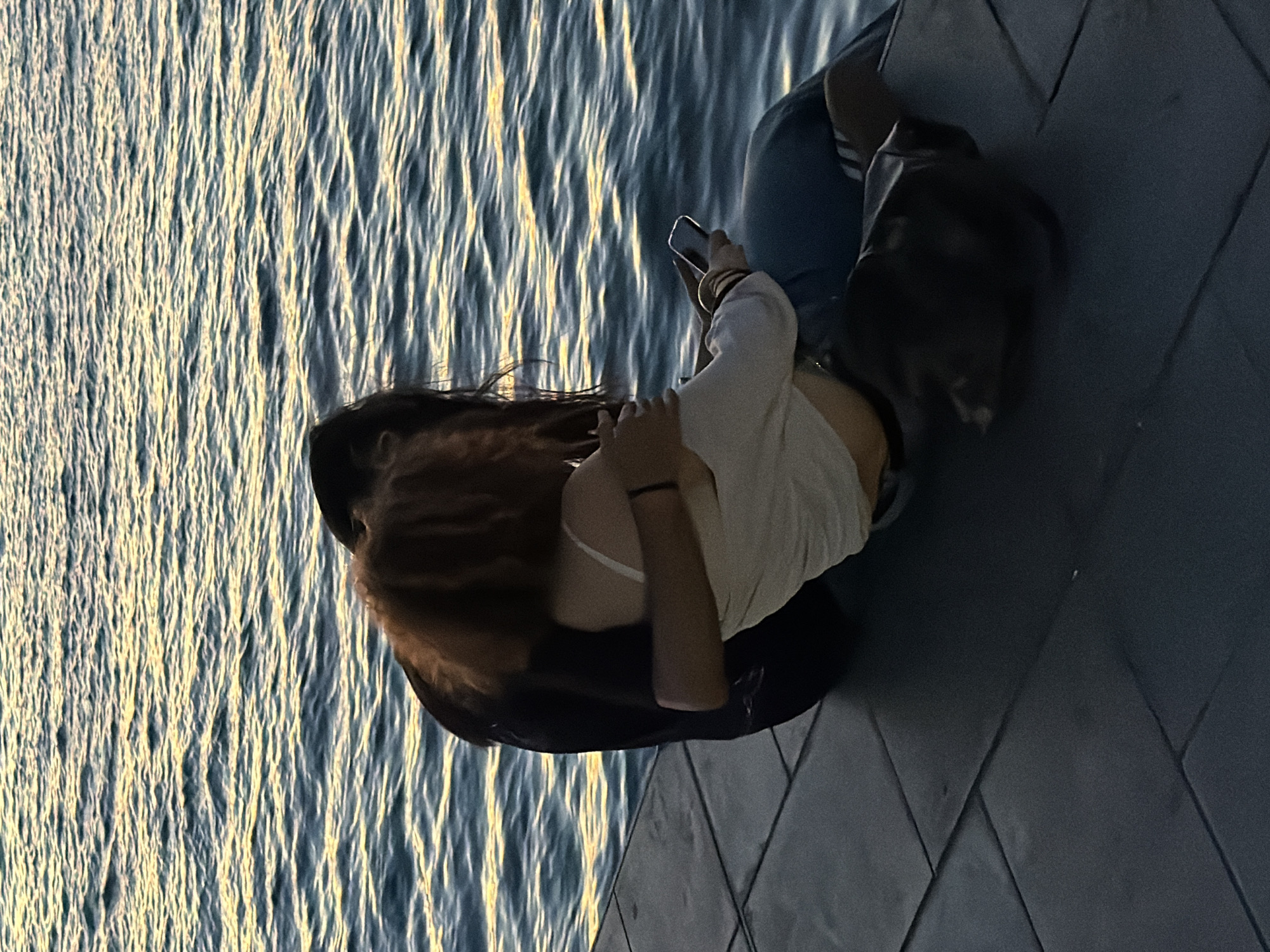 Two people are sitting on a tiled walkway next to the water in Kadıköy, Turkey. The person on the left, dressed in black, has their arm around the person on the right. The person on the right, with reddish-brown hair, is wearing a white tank top and blue pants, and is holding a smartphone in their right hand. The water behind them is rippling, reflecting the golden light of what appears to be sunset or sunrise. A dark bag is resting on the tiled surface between their legs.
