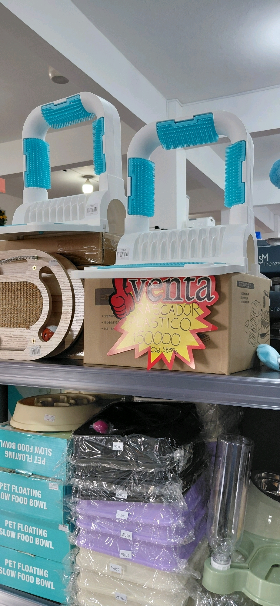 The image displays a retail environment, likely a pet supply store, in Encarnación, Paraguay. Two white U-shaped pet grooming or scratcher units with blue bristles are prominently featured on a shelf. To the right of these units, a cardboard box with a yellow and red "Venta" (Sale) sticker is visible, indicating a price of "150.000" and a product code "Cod. 38596". The text on the sticker appears to be in Spanish and potentially Portuguese, with words like "RASCADOR PLASTICO" (Plastic Scratcher) visible. Below this shelf, stacks of pet food bowls, identified by the text "PET FLOATING SLOW FOOD BOWL" on their packaging, are arranged. These bowls are in various colors including blue, black, purple, and white, with some displaying a price of "25000". A beige pet feeding station with a built-in water dispenser is also partially visible on the bottom shelf to the right. The background suggests an indoor commercial setting with white walls and ceiling lighting. The overall scene is well-lit and presents a typical retail display of pet products.