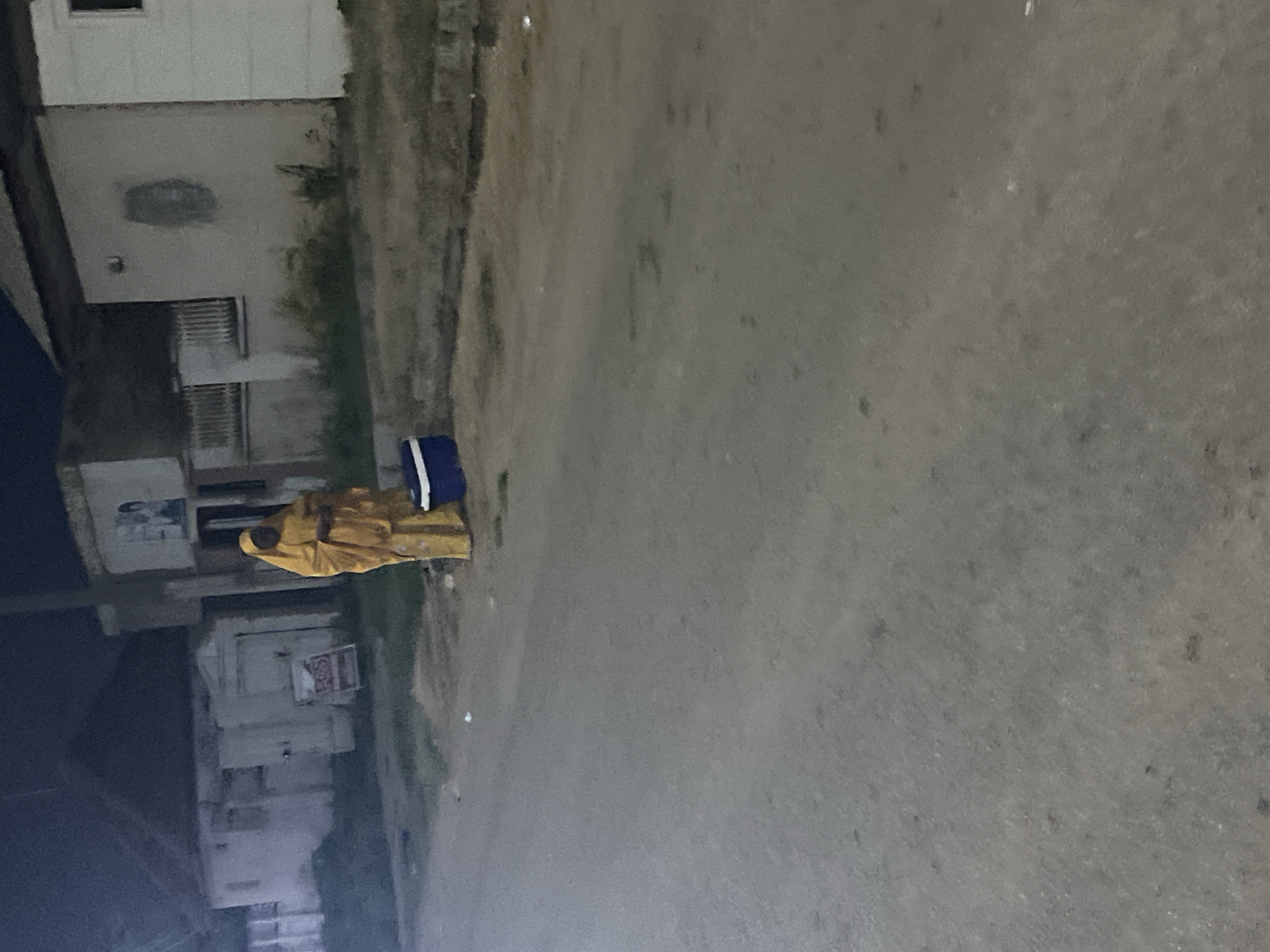 A person wearing a yellow hijab and long yellow garment stands on a paved street at night. Beside the person is a blue cooler. In the background are buildings with lights on. The scene appears to be in Abuja, Nigeria.