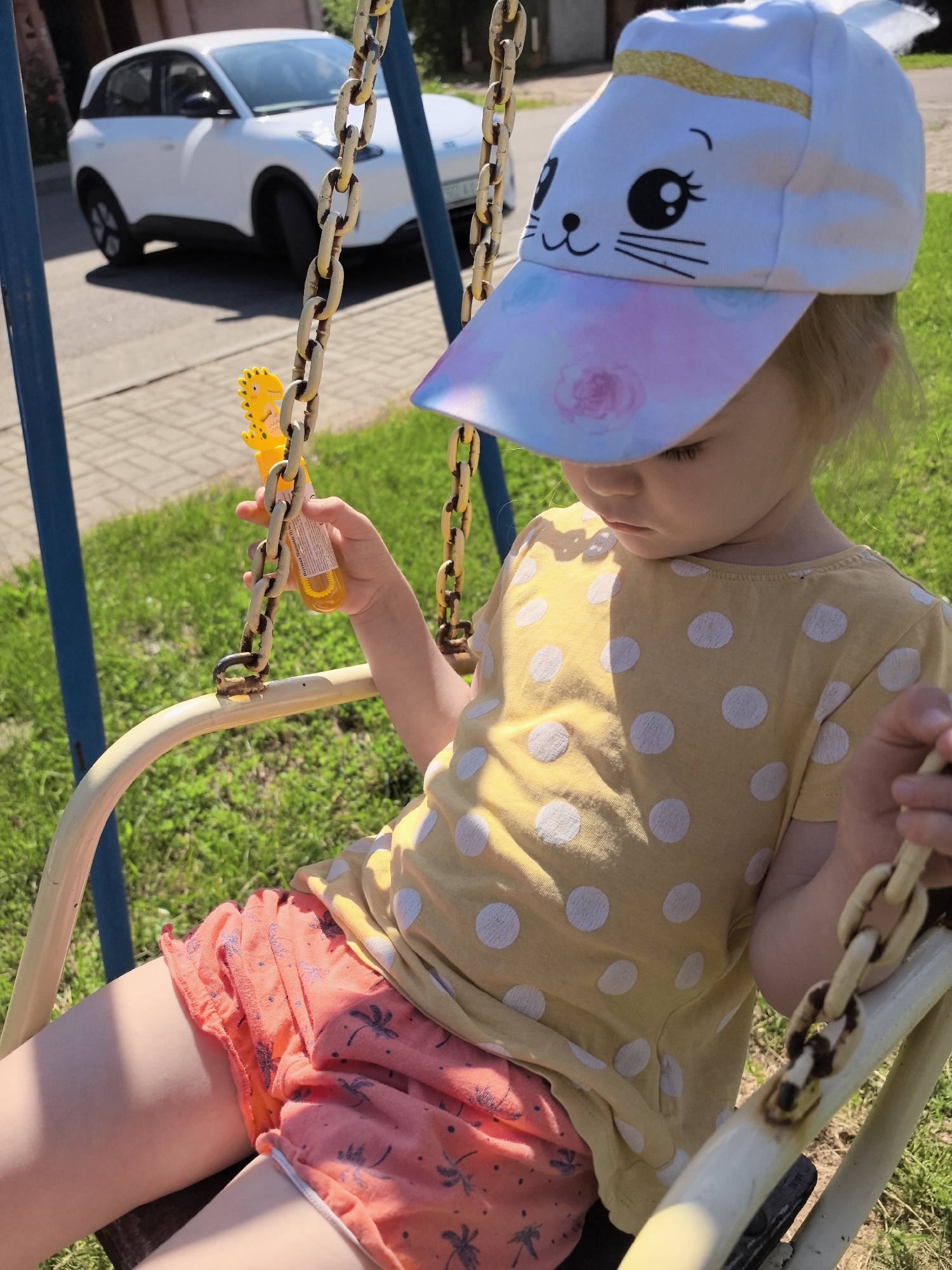A young child with blonde hair sits on a swing. She wears a yellow t-shirt with white polka dots and pink shorts with a palm tree pattern. On her head is a white baseball cap with a cartoon cat face and pastel-colored brim. Her left hand holds a small yellow bottle, possibly for blowing bubbles. The swing set has blue poles and rusty metal chains. Behind the child, a white SUV is parked on a paved surface. The background consists of green grass and some trees, suggesting an outdoor setting. The lighting indicates it's daytime, with bright sunlight casting shadows. There is no visible text other than on the bottle, which is too small to be legible.
