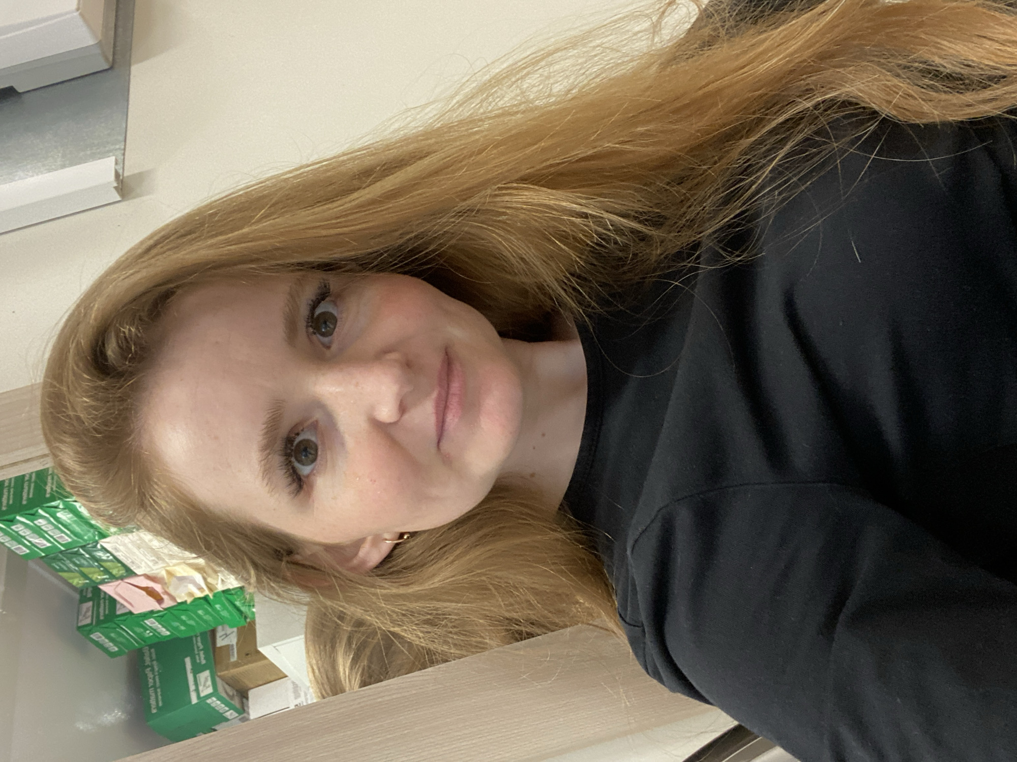 A young woman with long, light brown hair, fair skin, and light-colored eyes is depicted in an indoor setting. The image is rotated ninety degrees clockwise. She is wearing a black long-sleeved top and looks towards the camera with a calm, pleasant, and slightly smiling expression. Her hair flows around her face and over her shoulders.
The background shows stacks of green boxes, likely packaging for paper, with visible text including "premium", "500 sheets", and "80 g/m²". These boxes suggest an office, retail, or storage environment. Above the boxes, a light-colored wall and a part of a light fixture or ceiling structure are visible. A light brown wooden or laminate surface, possibly a desk or counter, is seen below her. The lighting is bright and even. The location context is Chaikovsky, Russia.