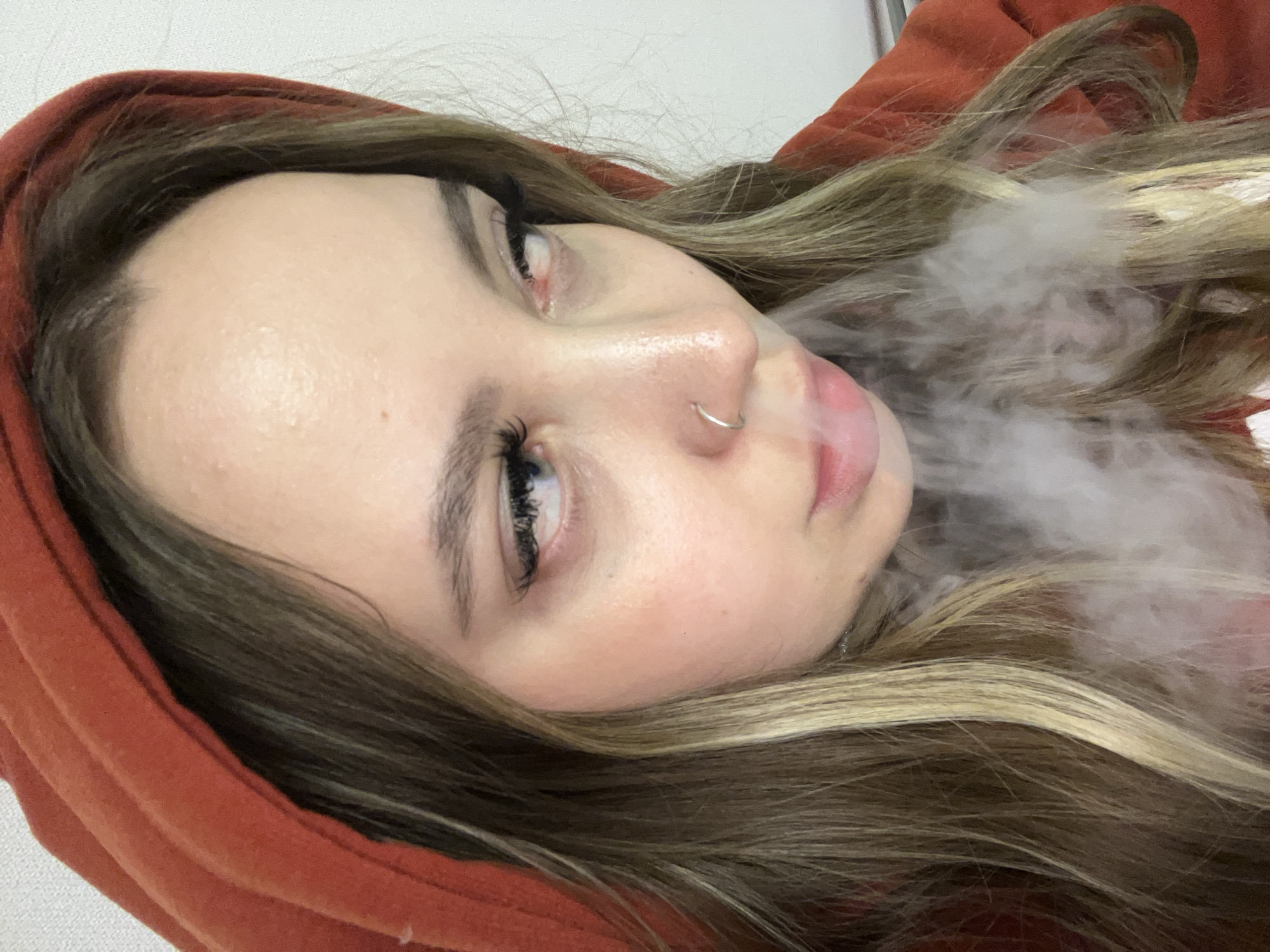 A person with light skin and brown hair is exhaling vapor or smoke. They are wearing an orange hooded garment. A silver ring is visible in the person's left nostril. The background appears to be a plain, light-colored wall. There is no information available to determine the location is Krasnodar, Russia.