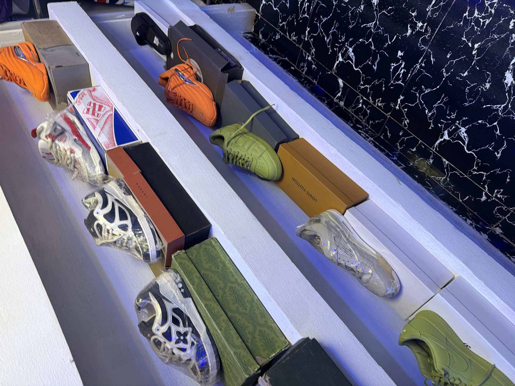 In Bukuru, Nigeria, a retail display features an assortment of athletic footwear. The shoes are presented on white shelves, with some items housed in their original boxes. The collection includes a pair of orange sneakers, a white and red sneaker in a box, and a black and white sneaker with a floral motif. Further down, a green sneaker rests on a brown box, and a clear-covered silver sneaker is positioned on a white box. A black slide sandal is also visible on a higher shelf. The floor is tiled with a black and white marble pattern.