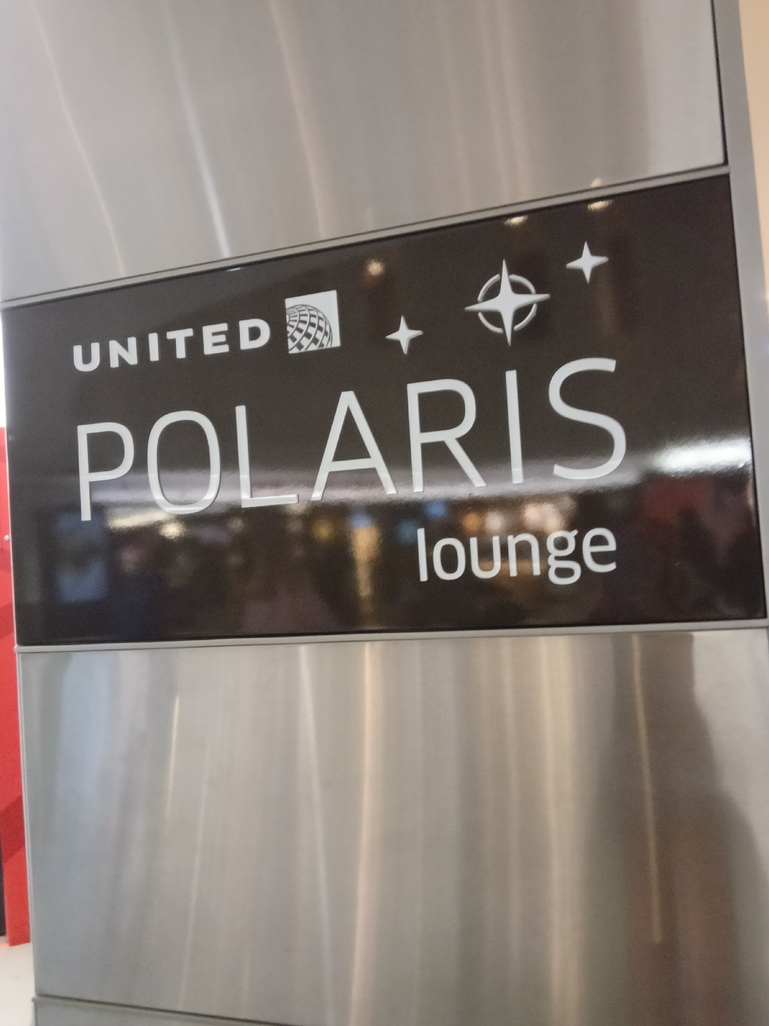 The image depicts a black rectangular sign for the "UNITED POLARIS lounge" set within a brushed silver-colored metal structure. This facility is located at Los Angeles International Airport (LAX), Los Angeles, California, United States of America.
The sign features white, sans-serif text: "UNITED" with its globe logo positioned to the right, followed below by "POLARIS," which is prominently displayed in larger font. To the right and slightly above "POLARIS," there is a white star/compass-like symbol, flanked by three smaller white four-pointed stars. Below "POLARIS," the word "lounge" is written in smaller white text. The sign's surface is glossy and reflective, showing blurry ambient lights and indistinct reflections of the surrounding airport environment. The metal paneling above and below the sign exhibits vertical brush marks and reflects ambient light, creating vertical streaks of varying brightness. No specific individuals are discernible within the frame. The scene is static, indicating an interior architectural feature within a public transportation hub.