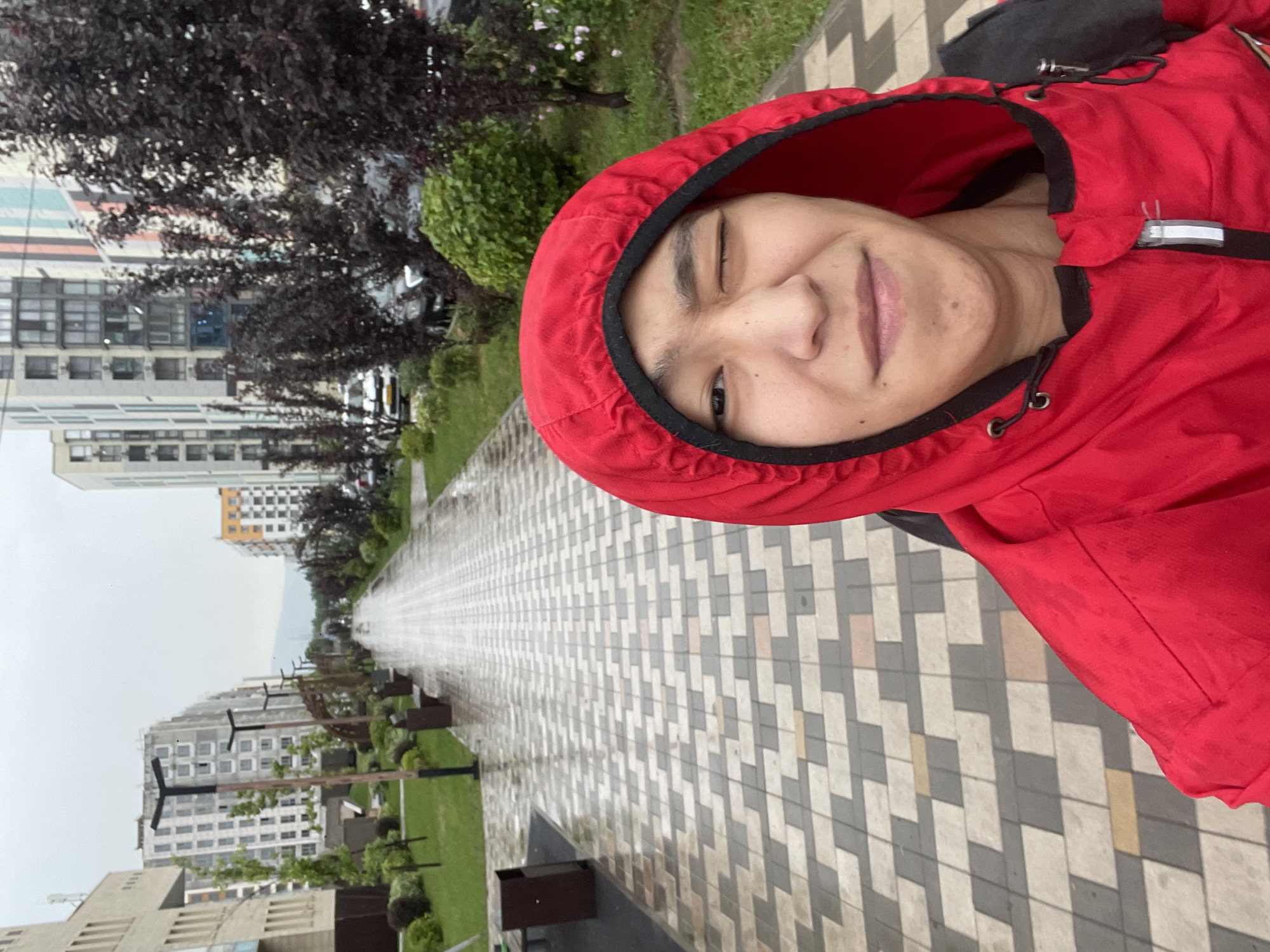 In Almaty, Kazakhstan, a young person wearing a red hooded jacket and backpack is taking a selfie on a wet, paved walkway. The walkway is lined with manicured green grass and decorative trees, with modern apartment buildings visible in the background. The sky is overcast, indicating a cloudy day. The person is winking at the camera, and the overall scene suggests a casual outdoor moment despite the weather.