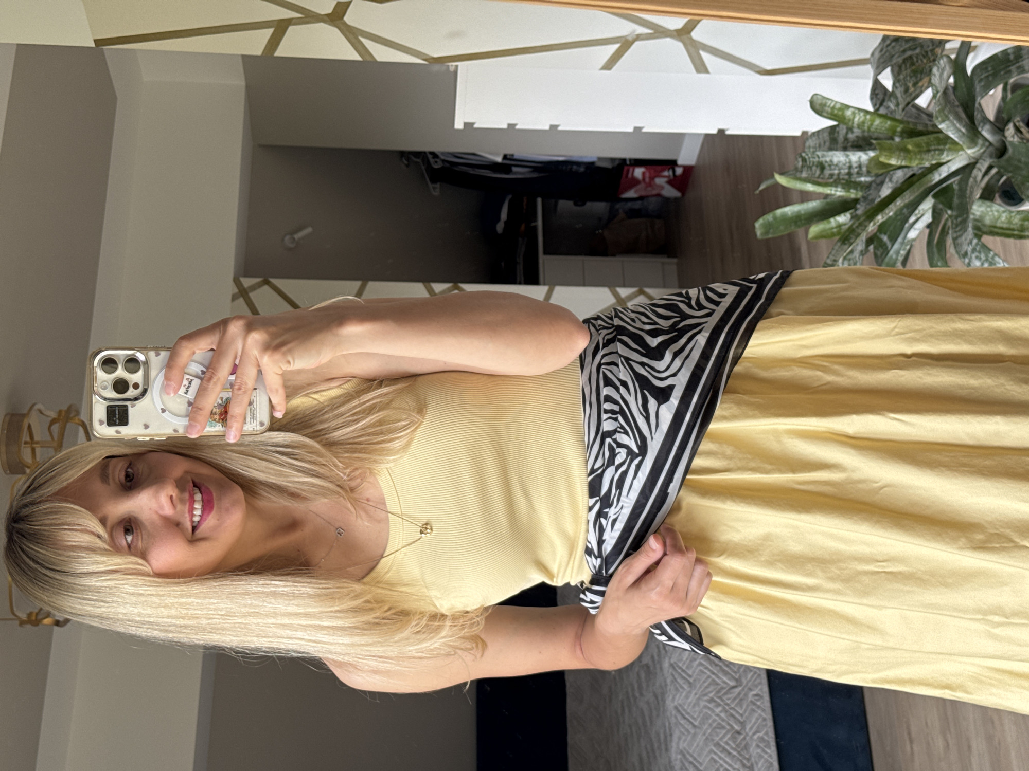 A woman with blonde hair and bangs, wearing a yellow tank top and a yellow skirt with a zebra-print scarf tied around her waist, takes a mirror selfie in a bedroom. The room appears to be in a residential setting, possibly an apartment in Carnikava, Latvia. In the background, a bed with a grey patterned comforter is visible, along with a closet with clothes. A potted plant sits to the right of the frame, and a mirror with a wooden frame is positioned prominently. The woman is holding a smartphone, which is decorated with stickers. The lighting suggests it is daytime.