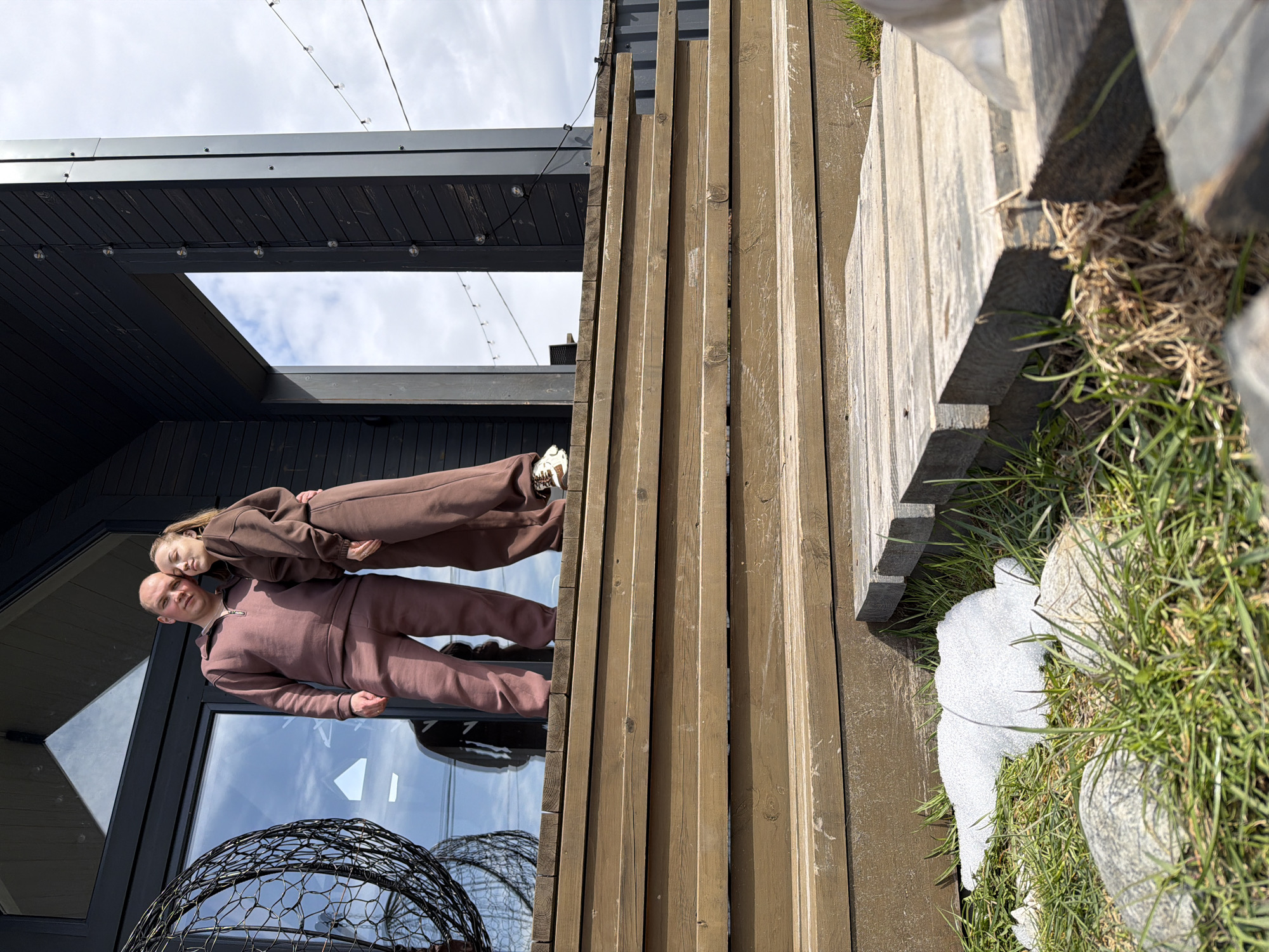 An outdoor, eye-level photograph, taken in Бунятино, Russia, captures two individuals standing beside a modern building on a bright, partly cloudy day. The original image is rotated 90 degrees counter-clockwise; described upright:
In the left-center, a man and a woman pose together. The man, bald and wearing a long-sleeved brown or dark mauve tracksuit with a quarter-zip collar, stands with his hands at his sides. The woman, with light brown hair, leans her head against his shoulder/back, also dressed in a matching brown tracksuit. Both look towards the camera with calm expressions.
They are positioned in front of a large, reflective window of a dark-colored, possibly black, modern building. The window reflects the bright sky and clouds. Below the window, a dark, open-weave metal sphere, possibly a decorative object or fire pit, is visible.
To the right of the people, a tall wooden slatted structure, appearing as a fence or part of the building's facade, dominates the right side of the frame. The wood is a natural, weathered light brown. Beyond this structure, in the foreground on the right, patches of green grass are visible, interspersed with melting snow, indicating an early spring or late winter season.
Above the building, the sky is visible with scattered white clouds. String lights with round bulbs are strung across the top of the frame, both attached to the building's dark roof structure and stretching across the open sky. Shadows suggest a clear light source, likely the sun. No visible text is present in the image.