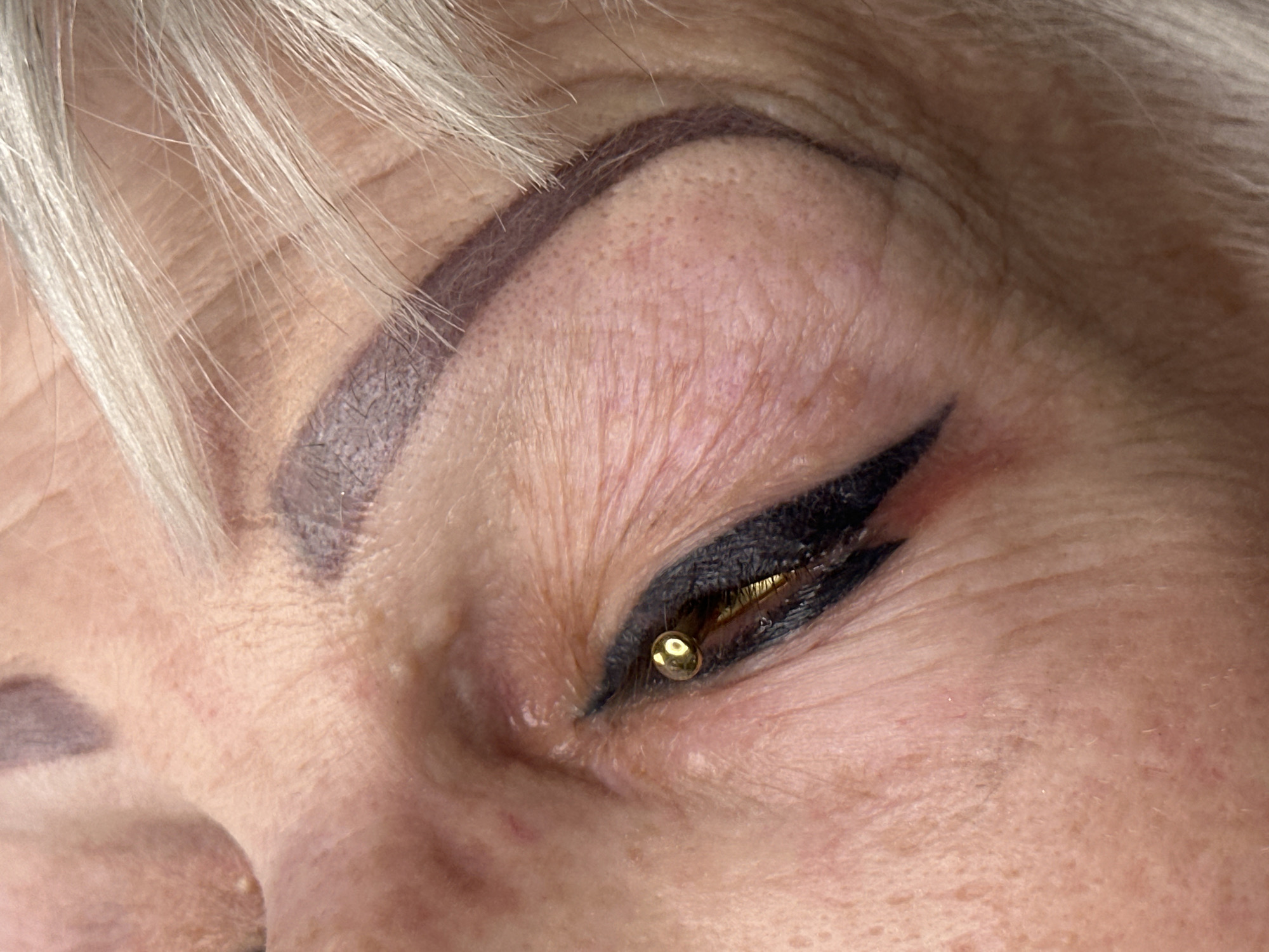 This close-up image focuses on the right eye and eyebrow area of an individual, likely female, showcasing cosmetic enhancements. The subject has light blonde or grey hair visible in the upper and lower left corners of the frame.
The eyebrow is precisely shaped and filled with a dark, solid color, resembling permanent makeup or a meticulously applied brow product. Fine lines and pores are visible on the skin around the eyebrow, indicating mature skin texture.
The eyelid features a dramatic, thick black winged eyeliner. A small, spherical, gold-colored object is prominently placed near the inner corner of the eye, resting on the lower lash line. This object, possibly a decorative piercing or an adhered gem, reflects light, adding a metallic highlight to the eye makeup. The eye itself is partially closed or looking downwards, revealing a glimpse of the dark eyeliner and the gold ornament.
The overall scene highlights detailed cosmetic artistry, possibly in a salon setting, emphasizing the precision of the eyebrow and eyeliner application, complemented by the unique eye adornment. No specific time of day, weather, or discernible emotions are conveyed. There is no visible text in the image. The broader context for this image is Riga, Latvia.
