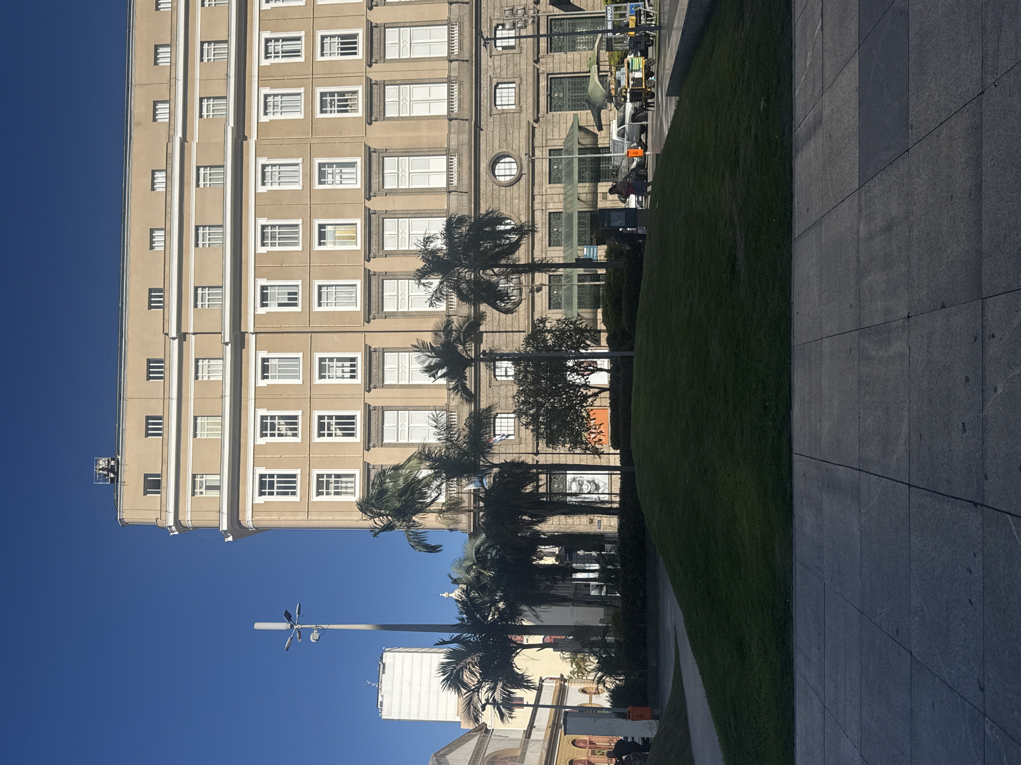 The image depicts an outdoor scene in the city of Rio de Janeiro, Brazil, likely near Cinelândia square, a historical and cultural hub. Dominating the frame is a large, beige, multi-story building with numerous white-framed windows, characteristic of the architecture in the area. In front of the building, a manicured, sloping green lawn is visible, leading towards a paved area in the foreground. Palm trees are scattered across the scene, some tall and slender, others shorter and fuller, adding a tropical element.
On the street level of the building, there are awnings and entrances. Several vehicles are present, including a white pickup truck, a small food cart with a green canopy, and other cars. People are sparsely distributed; a few individuals are walking on the sidewalk near the street, and one person is seated on the grass. A tall lamppost with multiple lights stands to the left of the main building, and further left, a taller, white building with a distinct antenna or structure on top is visible. The sky is a clear, bright blue, suggesting a sunny day. The overall impression is one of a public urban space, possibly a plaza or park area adjacent to significant architecture.
