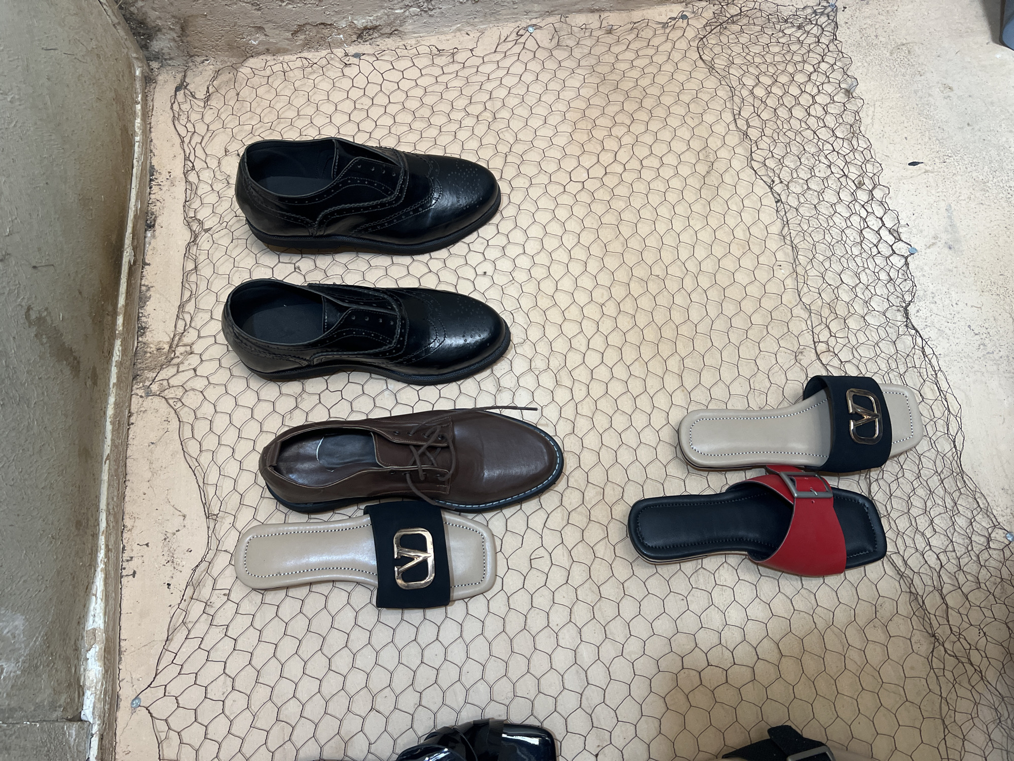The image depicts an assortment of footwear arranged on a surface covered with a wire mesh. The shoes consist of two pairs of black dress shoes, one pair of brown lace-up shoes, and two pairs of sandals, one with a black and beige color scheme and the other with a black and red color scheme. The setting appears to be indoors, potentially a retail space or storage area, located in Jos, Nigeria, as per the prompt.