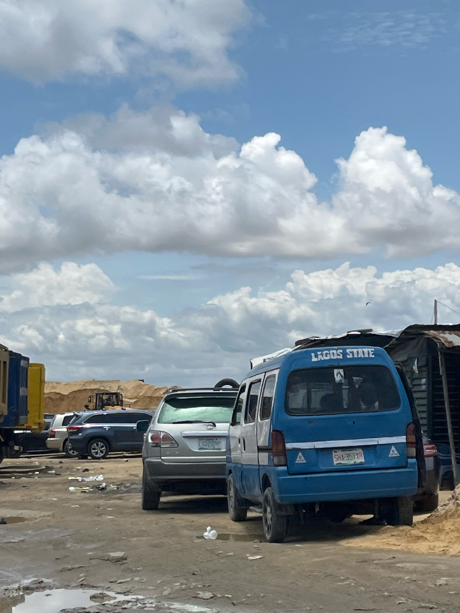 This is a wide shot, eye-level photograph taken outdoors on a cloudy day. The scene appears to be a dusty, possibly unpaved area with several vehicles. In the foreground, a blue and white minivan with "LAGOS STATE" written on the back is parked. To its left, a silver SUV is visible. Further to the left, part of a yellow and blue truck is in the frame. In the background, there are large piles of sand or dirt, with a bulldozer visible in the distance. There are also some makeshift structures on the right side of the image. The ground is uneven and appears to be muddy in some areas with scattered debris. The sky is filled with large, white cumulus clouds against a light blue backdrop. The overall impression is one of an industrial or construction site, likely in a developing area, given the presence of sand piles, heavy machinery, and informal structures. The vehicles suggest a location within or related to Lagos State, Nigeria.