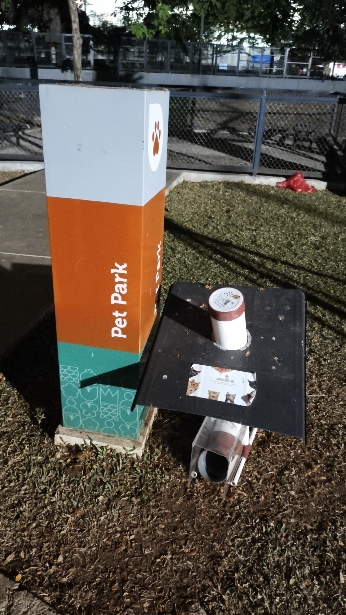 The image captures a pet park amenity in South Jakarta, Indonesia. The primary subject is a tall, three-sectioned post with a grey top section featuring a paw print graphic, an orange middle section with the text "Pet Park" in white, and a teal bottom section adorned with white line drawings of various animal-related icons such as butterflies, crowns, and paw prints. Adjacent to the post, a black, angled structure is positioned, serving as a dispenser or collection point. This structure has a white tube with a brown cap extending upwards, and a transparent tube at the bottom, possibly for waste disposal. A sticker depicting several cats is adhered to the black surface of the dispenser. The scene is set outdoors, with dry grass and fallen leaves covering the ground. In the background, a chain-link fence encloses an area with trees and some industrial-looking structures, suggesting an urban park or recreational space. A red plastic bag is visible on the grass near the fence. The lighting suggests it is either early morning or late evening.
