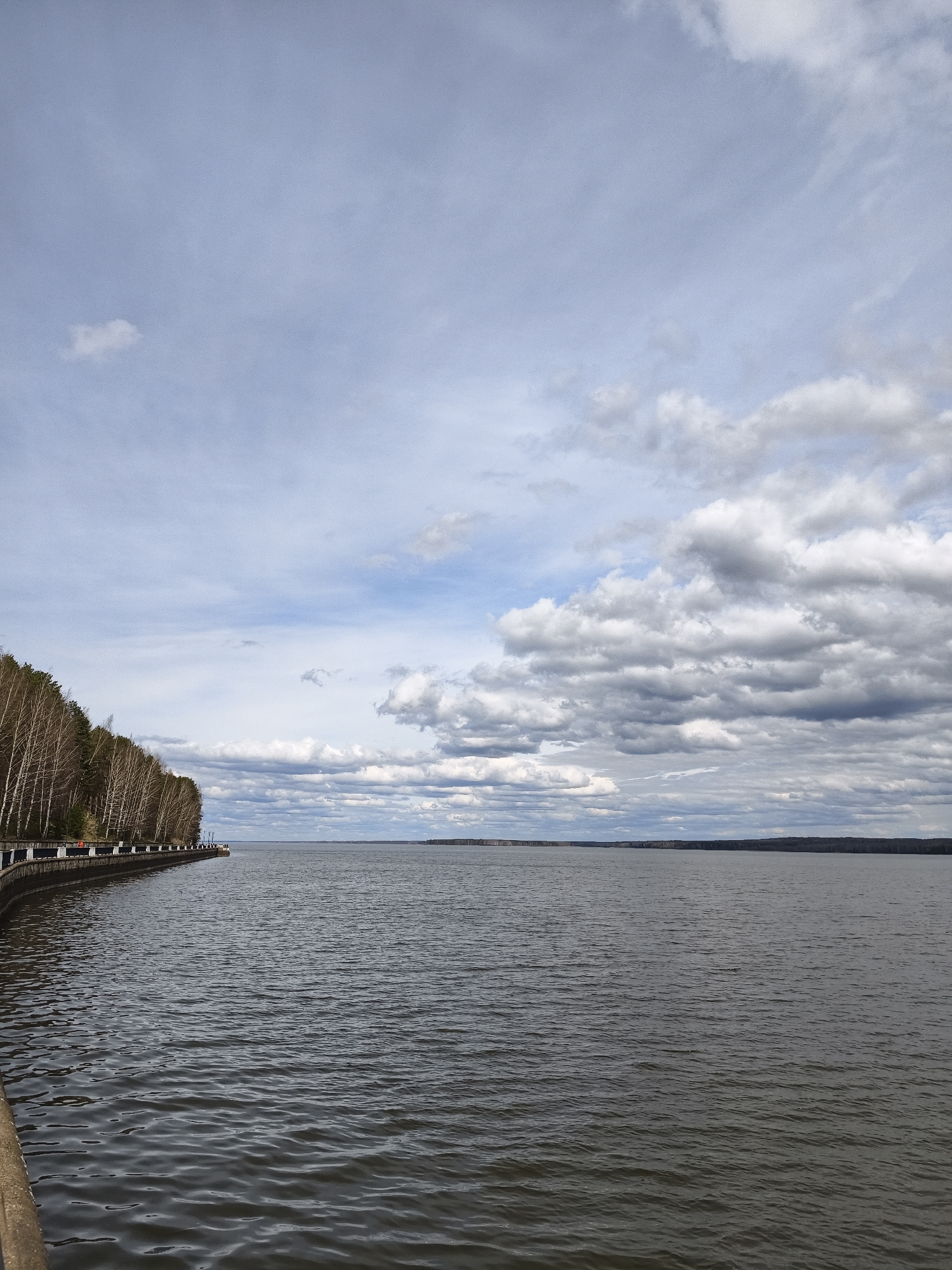 The image depicts a wide expanse of water, likely a large river or reservoir, under a cloudy sky. The location is Chaikovsky, Russia. On the left side of the frame, a paved embankment runs along the water's edge, lined with a row of trees. The trees appear to be deciduous, with bare branches, suggesting it is either late autumn, winter, or early spring. In the distance, across the water, lies a low-lying landmass, potentially an island or the far shore of the water body. The sky is filled with clouds of varying density, some light and wispy, others darker and more ominous. The water surface is slightly rippled, indicating a gentle breeze. No people are visible in the image.