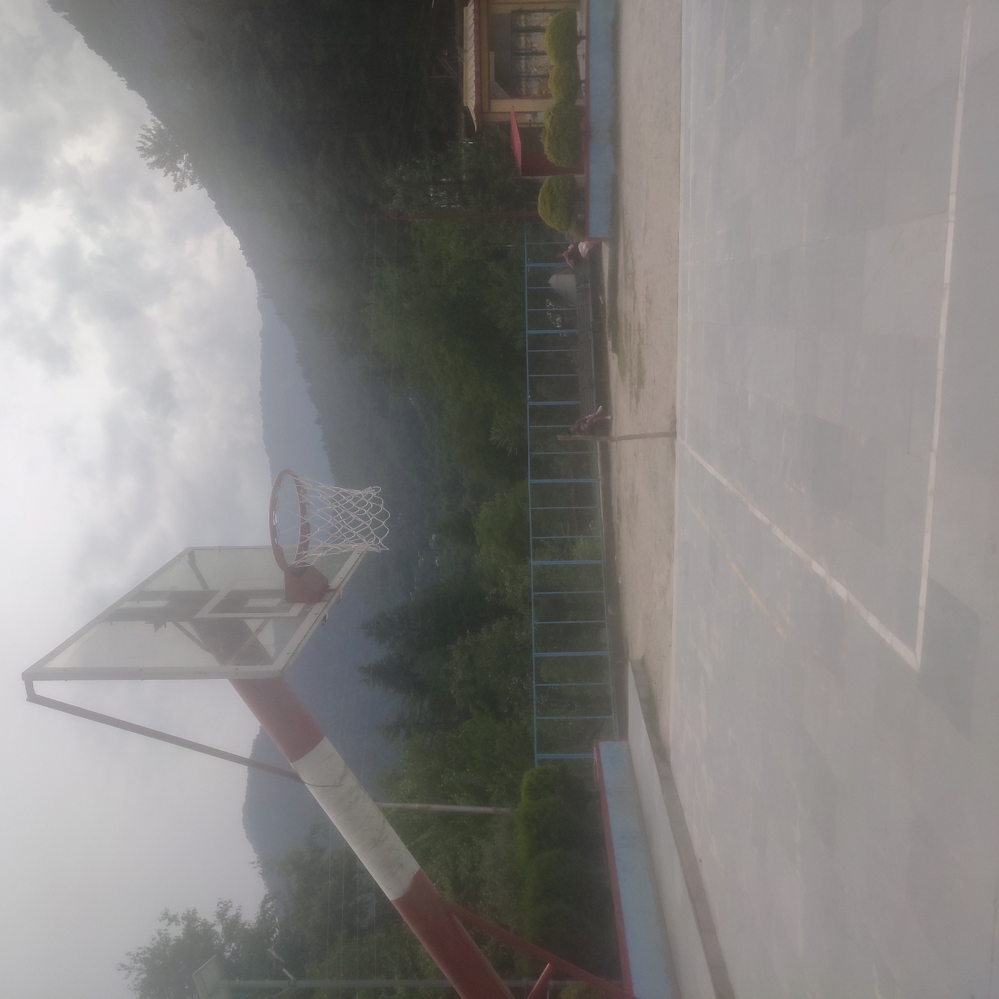 The image depicts an outdoor basketball court situated in Batahar, India, under a hazy or cloudy sky. In the foreground, the court surface is visible with white markings. A basketball hoop with a white net is prominently featured on the left side of the image, extending from a red and white pole. The court is bordered by a low concrete wall with a blue trim. Behind the court, a blue metal fence runs horizontally. Further in the background, there are several individuals seated or standing, some near a bench. Beyond the fence, dense green foliage and trees are visible, leading up to a mountainous landscape shrouded in mist. A building with a distinctive roof structure and ornate detailing is visible on the right side of the background, possibly a temple or cultural center. The overall impression is of a serene, possibly humid, environment with a backdrop of natural beauty.