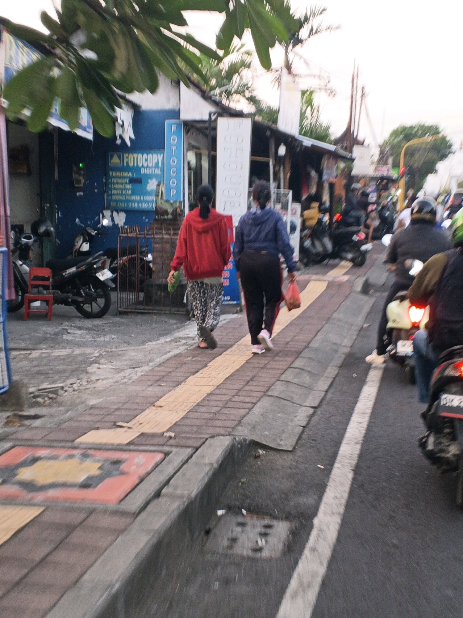 The image depicts a street scene in Denpasar, Indonesia, likely during the day with overcast skies. The foreground shows a textured sidewalk with a drain. On the left side of the sidewalk, a red plastic chair sits beside a parked motorcycle. Behind the chair and motorcycle, a building with a blue facade is visible, featuring a sign for "FOTOCOPY DIGITAL" and a green indicator light.
Moving towards the center and right of the image, two women are walking on the sidewalk. The woman on the left wears a red hooded jacket and a patterned skirt, carrying a green object in her right hand. The woman on the right wears a dark blue jacket and black pants, holding a reddish-brown plastic bag in her right hand.
The street beside the sidewalk is occupied by a number of motorcycles and riders. Several motorcycles are seen in motion, with their brake lights illuminated. One rider in the foreground is wearing a black helmet. The street is paved and has a white dividing line. Buildings with various awnings and signs line the right side of the street, suggesting a commercial area. Tropical foliage is visible in the upper left portion of the image, and utility poles are present in the background. The overall impression is that of a busy urban street with pedestrian and vehicular traffic.