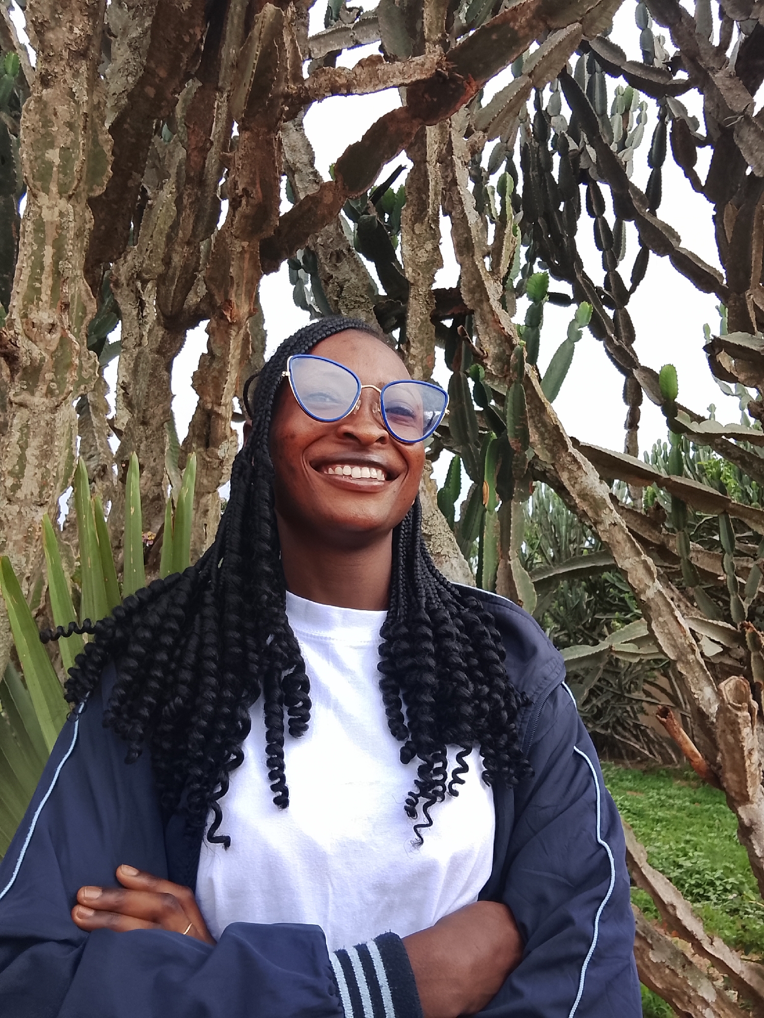 A young Black woman with dark braided hair and blue-framed glasses is standing with her arms crossed, smiling broadly. She is wearing a white T-shirt and a navy blue jacket with light blue stripes on the sleeves. The background consists of tall, thick cacti of varying shapes and sizes, interspersed with some green foliage and a patch of grass. The sky is overcast. The location is likely in Kuru Dungus, Nigeria, based on the prompt instructions.