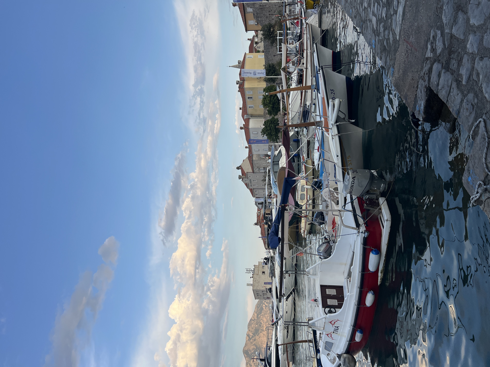 The image depicts a harbor in Budva, Montenegro, with numerous boats docked along the waterfront. The sky is blue with scattered white clouds, suggesting it is daytime. In the background, buildings with red-tiled roofs line the shore, with a distant mountain range visible. The foreground shows several boats of various sizes, some appearing to be small motorboats and others larger yachts. The water in the harbor is calm, reflecting the sky and the boats. A stone walkway or pier is visible along the right side of the image. No people are clearly identifiable in the scene.
