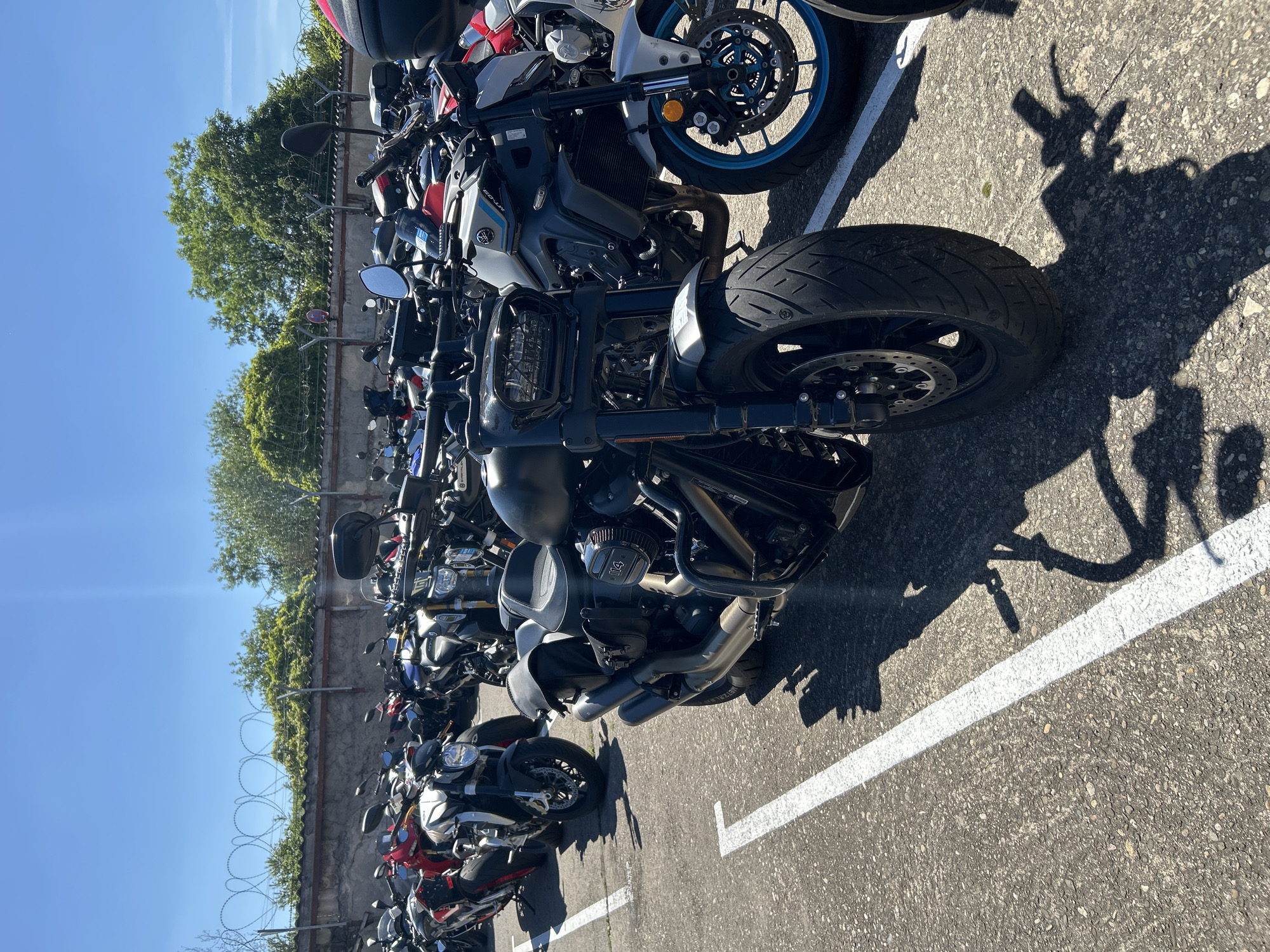 The image shows a row of parked motorcycles in an outdoor lot, possibly in Bernhausen, Germany (as indicated in the prompt). The motorcycles are various makes and models. A tall wall with barbed wire runs along the edge of the parking lot, beyond which are trees. The sky is clear and blue. The parking lot is marked with white lines. The motorcycles cast long shadows, suggesting it is midday.