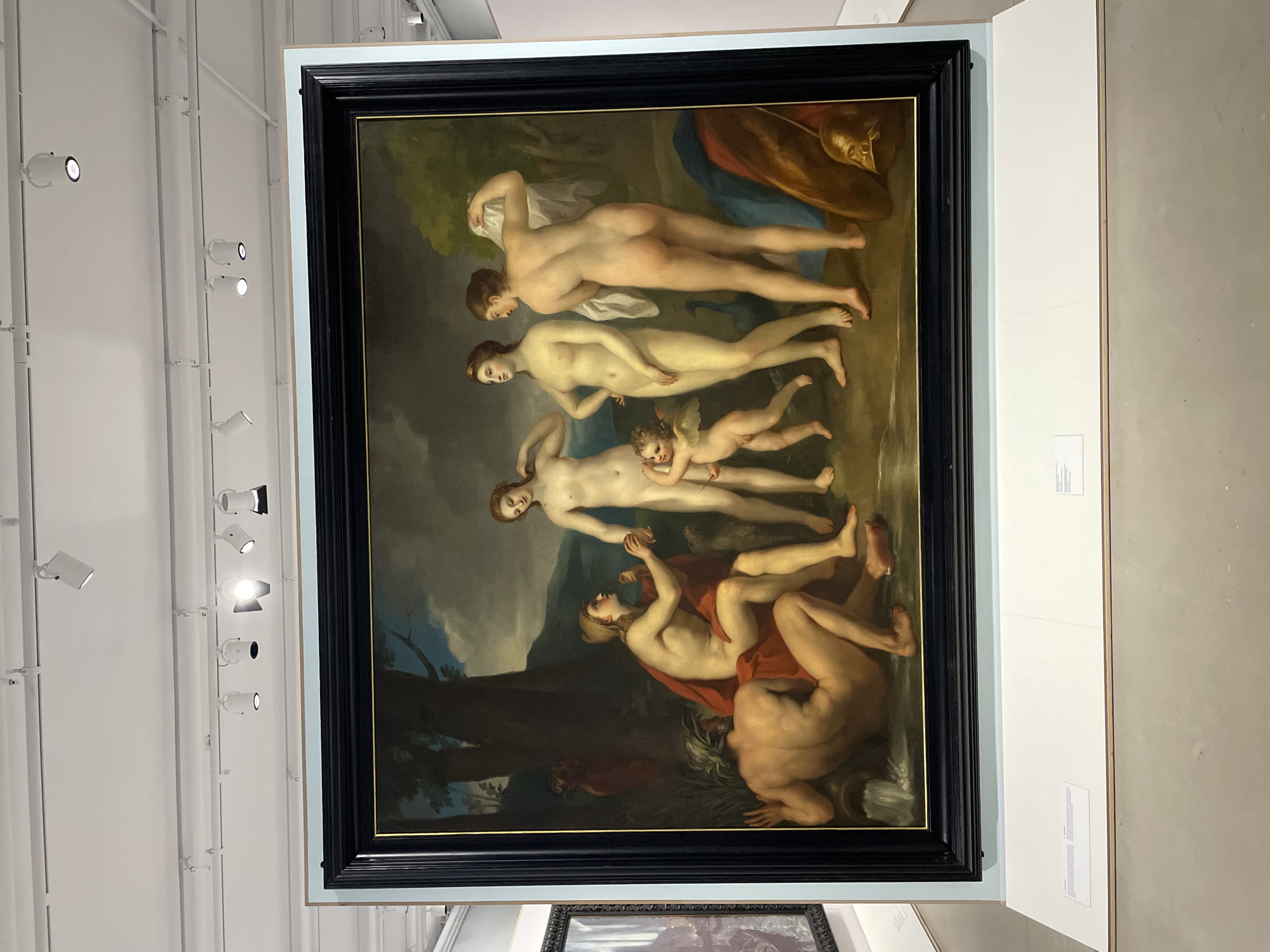 The image depicts a framed painting on display in a museum or gallery setting. The painting itself features a classical or mythological scene with nude figures, including women, a child with wings (possibly Cupid), and a reclining male figure. The figures are arranged in a composition against a landscape backdrop with trees and clouds. The painting is framed in a dark, ornate frame. The surrounding environment shows museum walls, lighting fixtures, and possibly other artwork or displays in the background. There are no people visible in the image outside of the painting itself. The location of the image is in Moscow, Russia.