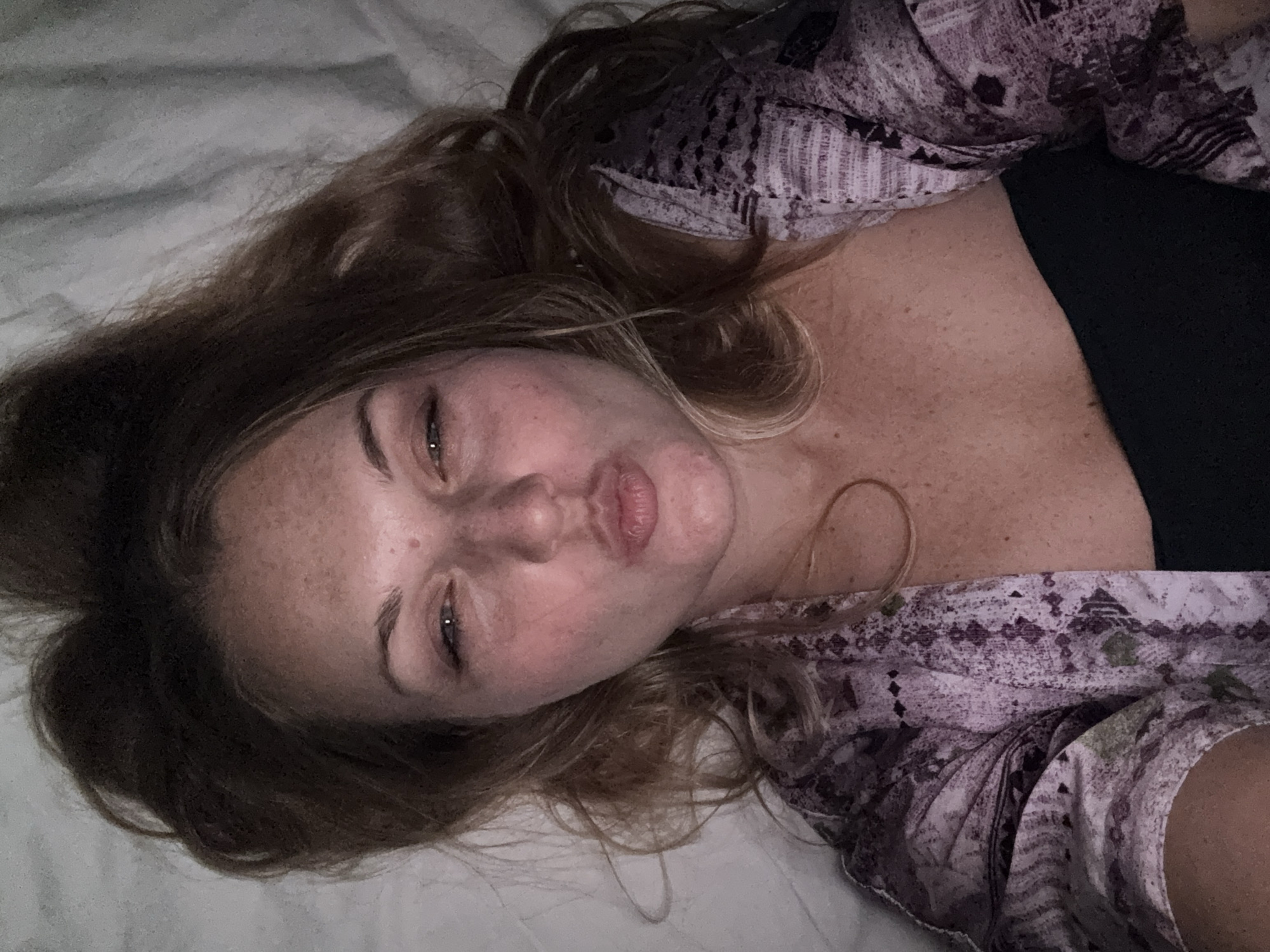 A woman lies in bed, facing the camera and puckering her lips. She has long, messy brown hair and freckles across her face. She is wearing a purple and pink patterned robe over a black top. The background is a white, wrinkled sheet. There is no indication of the city of Denpasar, Indonesia, in this image.