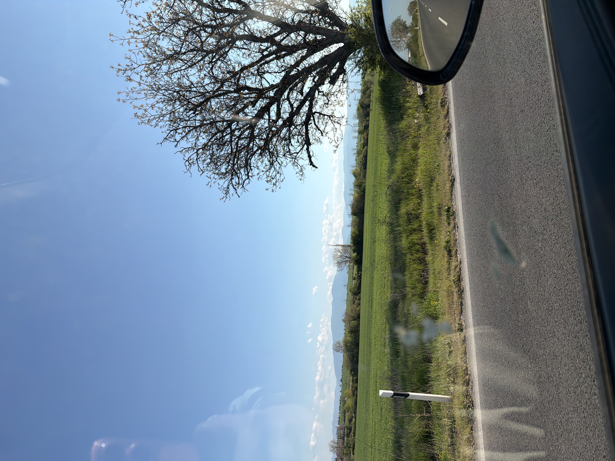 The image shows a view from a car window on a sunny day. The scene includes a road, a green field, a tree with bare branches, and a clear blue sky. A side mirror reflects the road ahead. A white post with a black stripe is visible along the side of the road. There are mountains on the horizon. The location is Unknown, Unknown.
