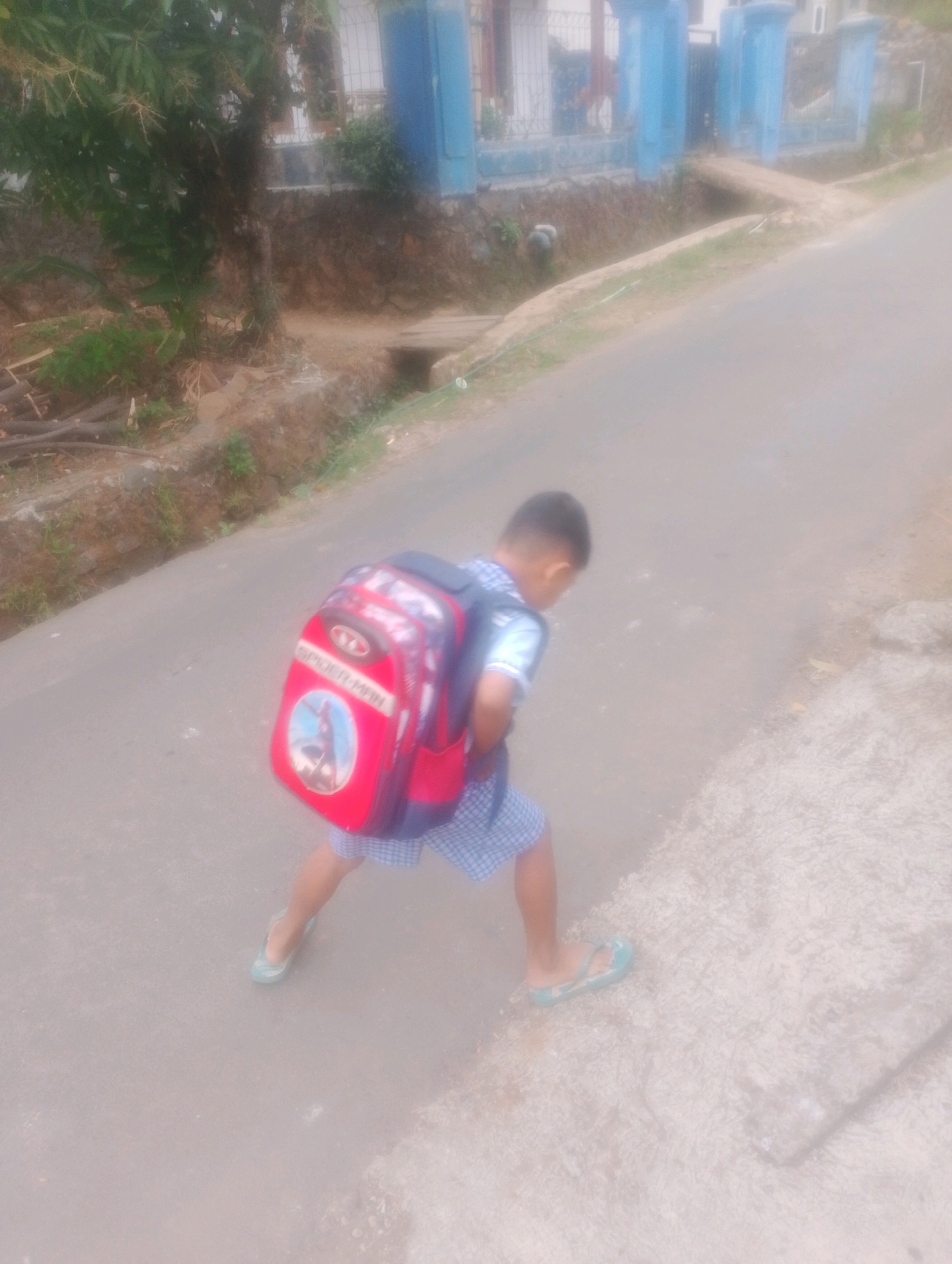 In Kalapadua, Indonesia, a young boy with a Spiderman backpack walks on a paved road. He is wearing a white t-shirt with blue trim and blue and white checkered shorts, along with light blue flip-flops. The road is bordered by a sloping, vegetated embankment on the left and a textured surface on the right. In the background, a blue fence and building are visible.