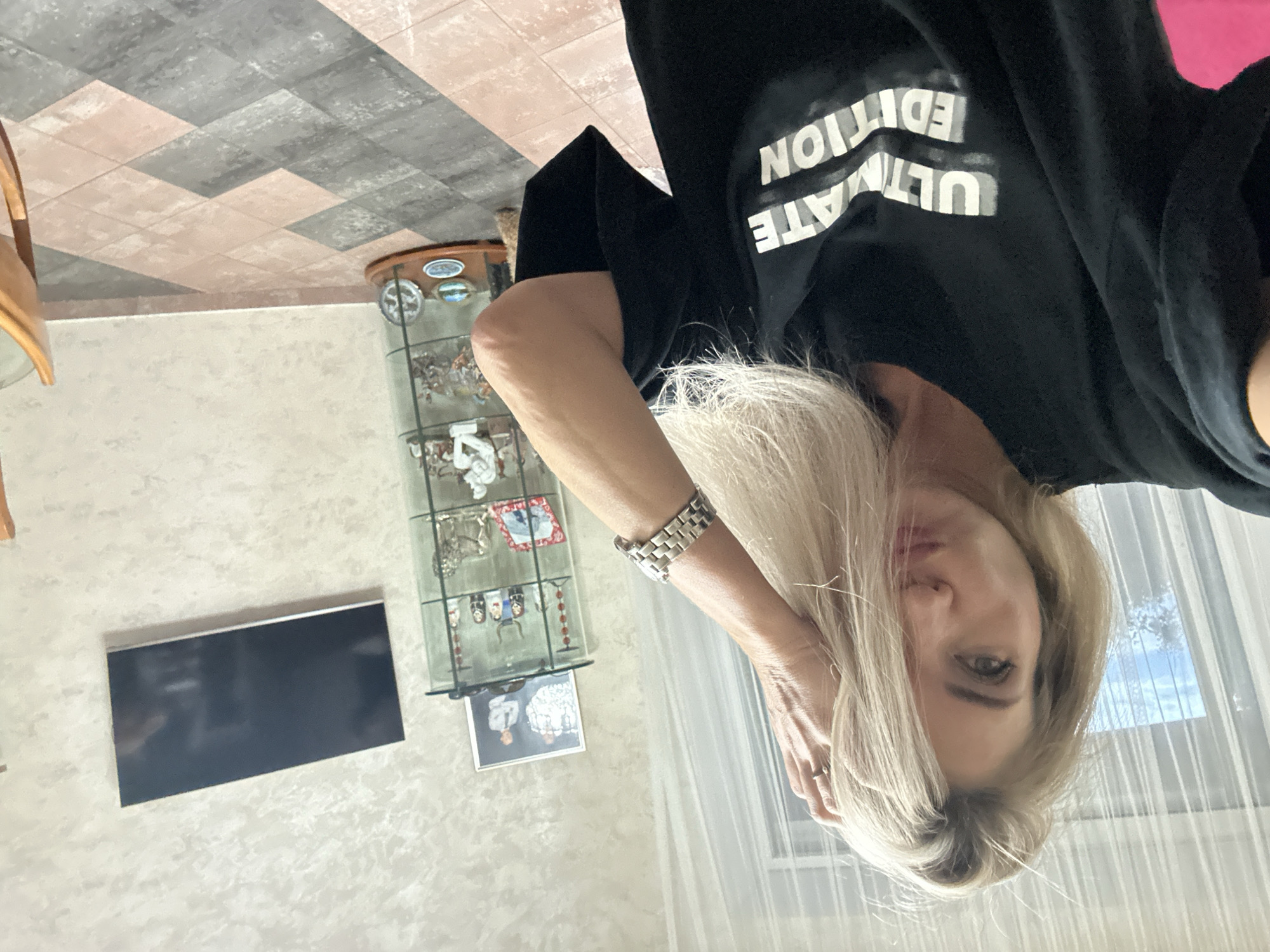 The image is a selfie of a woman with blonde hair, wearing a black t-shirt with white text that reads "ULTIMATE EDITION". She is posing with her hand in her hair and her head tilted. In the background, there is a display cabinet filled with figurines, a framed photograph of two men, and a flat-screen television mounted on the wall. The room has textured wallpaper and a tiled floor. The scene appears to be an indoor residential setting. There is no specific information about the city of Armavir, Russia, in the image.