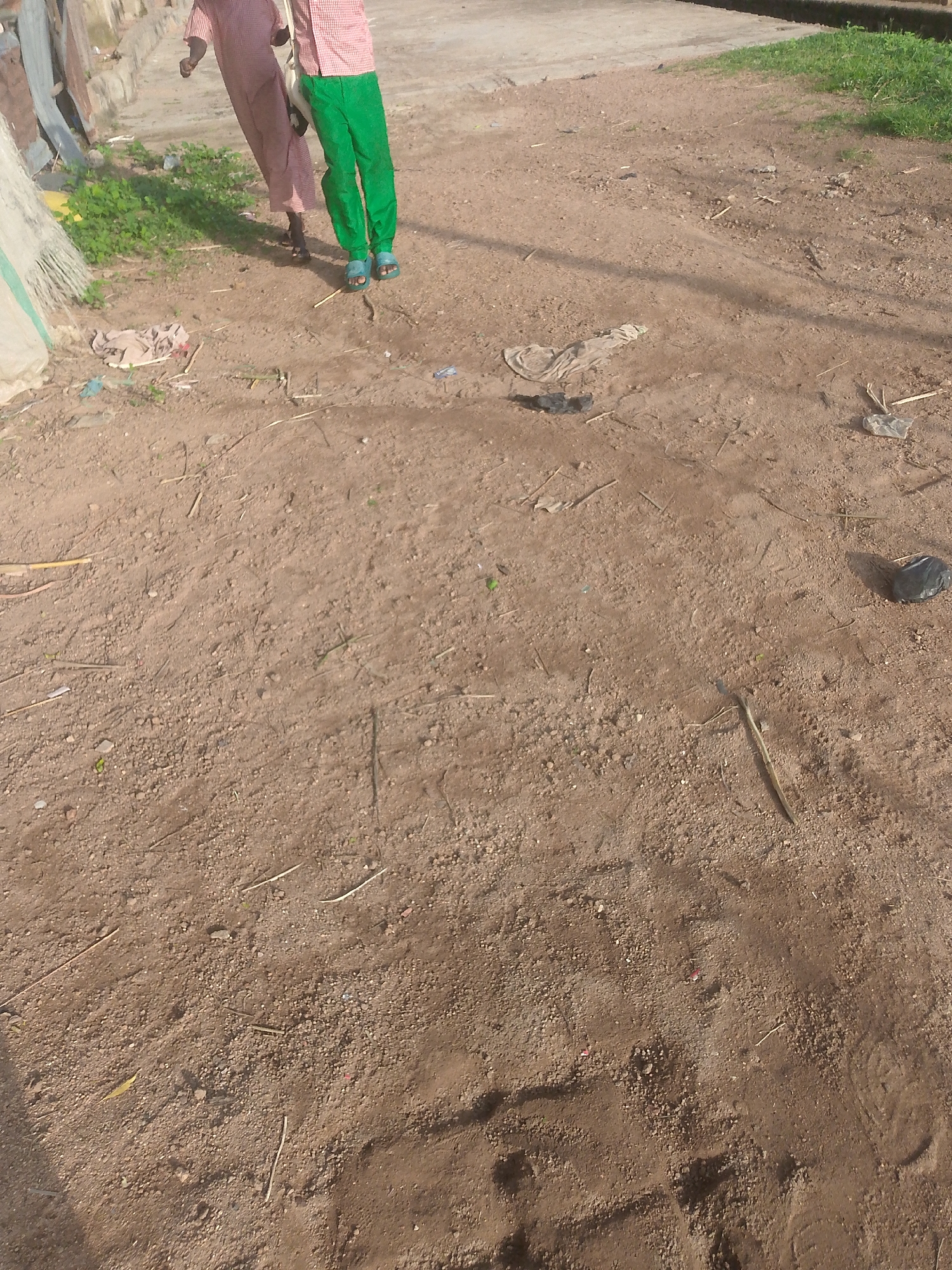 In Jalingo, Nigeria, two individuals stand on a dirt path. One person, on the left, wears a long, pink, checkered garment and dark sandals. The other person, to the right, has on a red and white checkered shirt and vibrant green pants, with blue sandals. The ground is composed of dry, brown earth, interspersed with small stones and scattered debris including twigs, plastic bags, and fabric scraps. To the left of the path, green foliage grows abundantly. In the background, a concrete structure and more vegetation are visible. The scene appears to be an outdoor setting, possibly a residential area or a less-maintained public space.