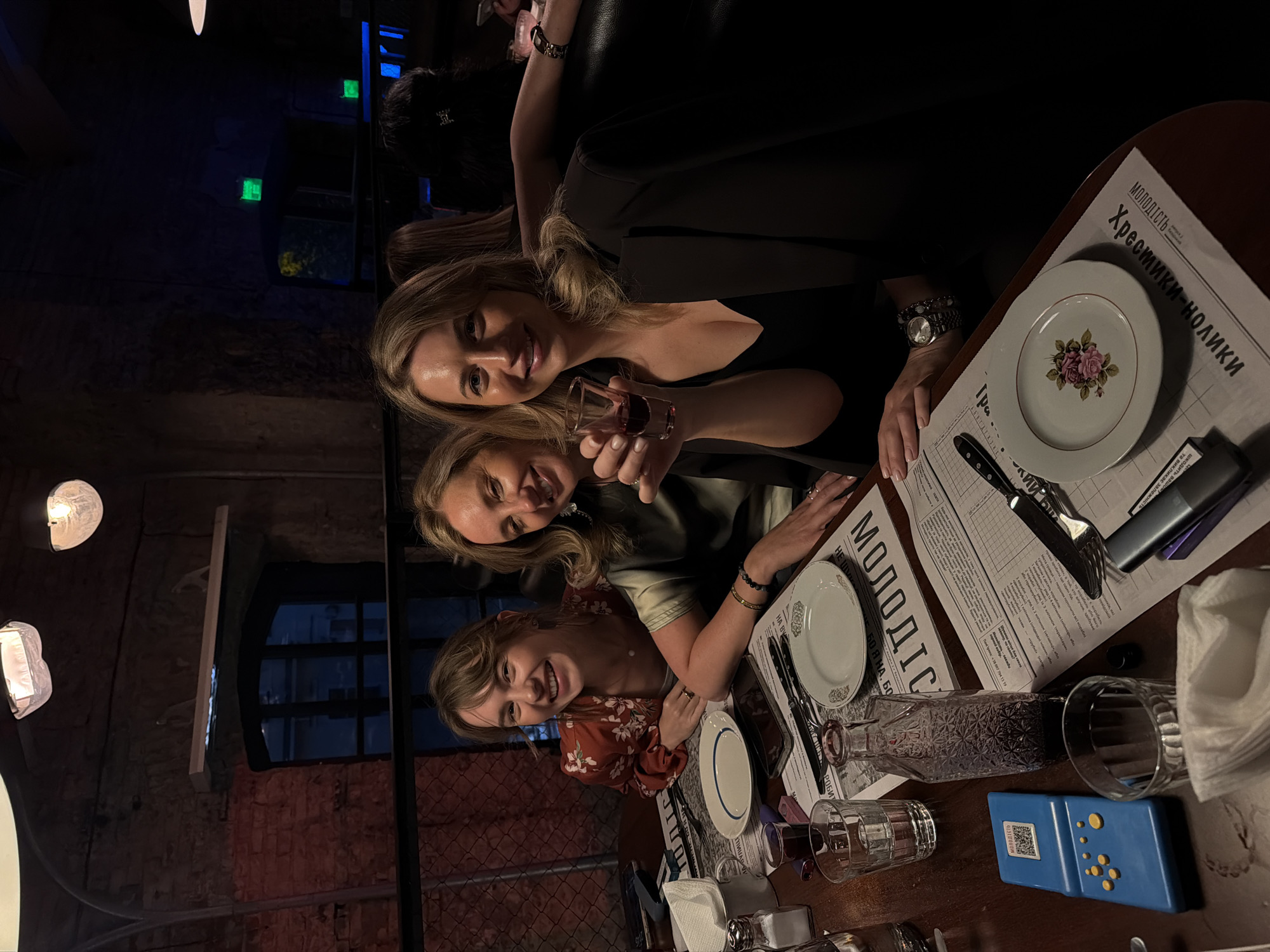 The image shows three women seated at a table in a dimly lit restaurant. The woman on the left is smiling and wearing a floral-patterned top. The woman in the middle is wearing a dark green top and holding a small shot glass filled with a dark liquid. The woman on the right is wearing a dark blazer over a black top and is also smiling. The table is covered with menus and place settings, including plates, glasses, and silverware. A blue electronic device and a glass bottle are also visible on the table. The background suggests an industrial-chic interior with exposed brick walls and hanging light fixtures. The overall impression is of a social gathering. While the image doesn't explicitly show the location as Kyiv, Ukraine, the presence of Cyrillic text on the menus suggests a Slavic-speaking region.