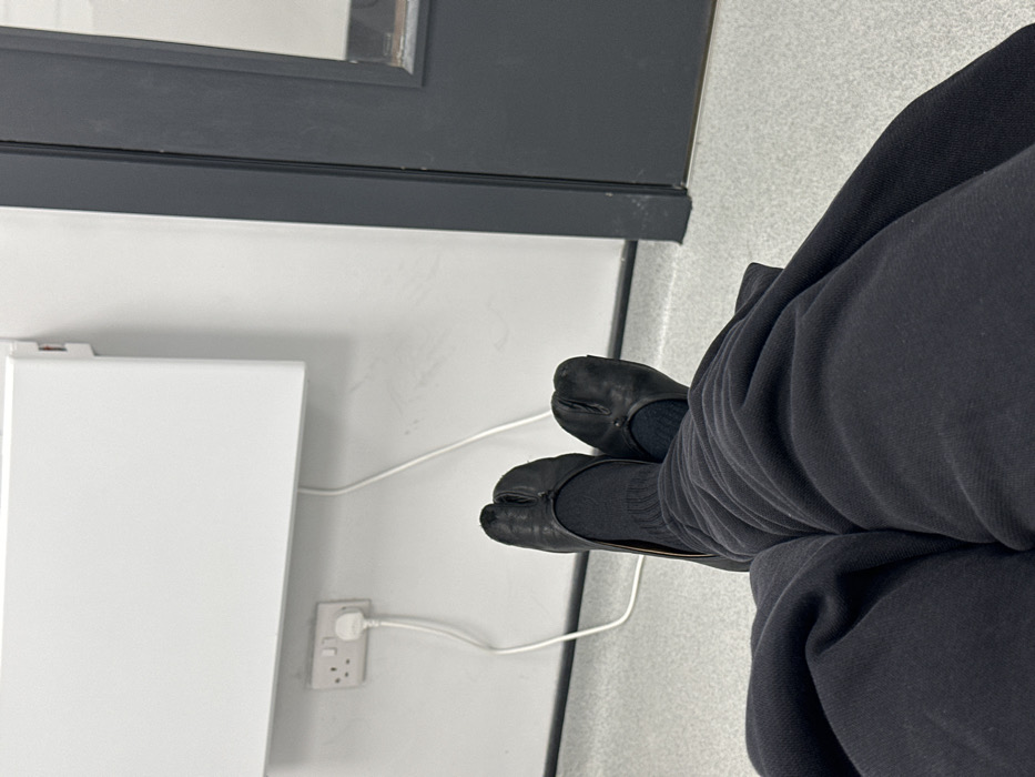 The image displays a close-up, downward perspective of a person's lower legs and feet, suggesting they are seated in an indoor environment. The person wears black, seemingly comfortable, trousers or sweatpants and black ribbed socks. Their feet are adorned with distinctive black tabi-style shoes, which are flats or loafers characterized by a split-toe design.
The setting includes a plain white wall on the left, against which a white rectangular appliance is mounted. This appliance could be a panel heater or a small storage unit. Below and to the right of the appliance, a white UK-style triple gang power outlet is visible, with a white plug inserted into one of its sockets. A white electrical cable extends from this plug, running along the wall and floor. The floor is light grey, possibly tiled or carpeted. In the background, a dark grey or black horizontal structure, likely the bottom frame of a door or a window, is visible.
The overall scene suggests a functional or minimalist interior space, possibly a waiting area or an office. Given the location context, this is in Tunbridge Wells, United Kingdom, consistent with the UK-style power outlet. No specific activity beyond sitting is evident, and there are no discernible emotions, time of day, weather conditions, or visible text.