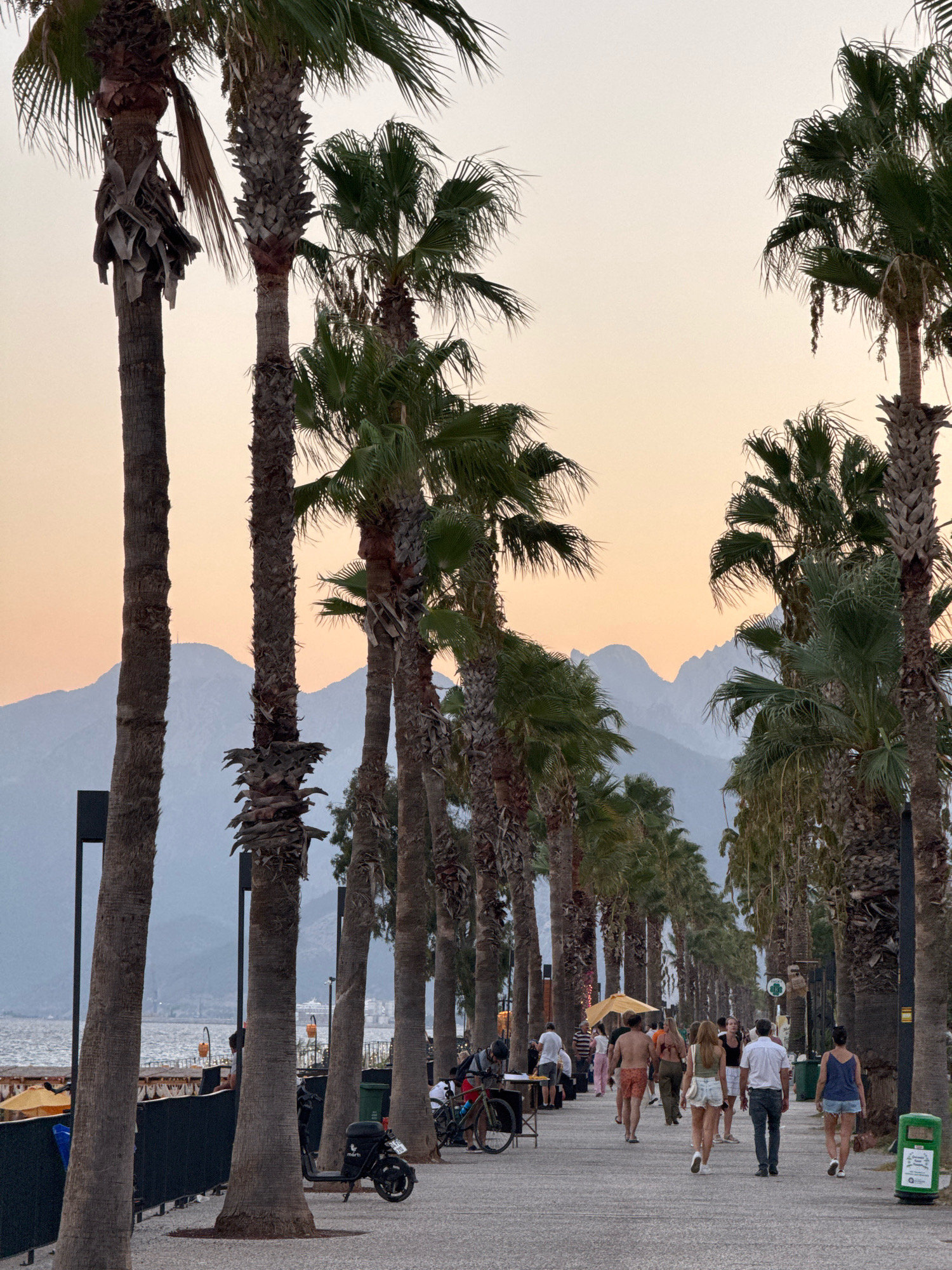 The image depicts a scenic promenade in Konyaaltı, Turkey, lined with numerous palm trees. The promenade is paved and features a walkway where several individuals are seen strolling. The background is dominated by a hazy mountain range under a soft, dusky sky. On the left side of the promenade, a body of water can be glimpsed, with a breakwater and some distant buildings. There are also several black lampposts and a black electric scooter parked on the walkway. Several people are dressed in casual summer attire, suggesting a warm climate. The overall atmosphere is peaceful and leisurely, typical of a seaside resort town.