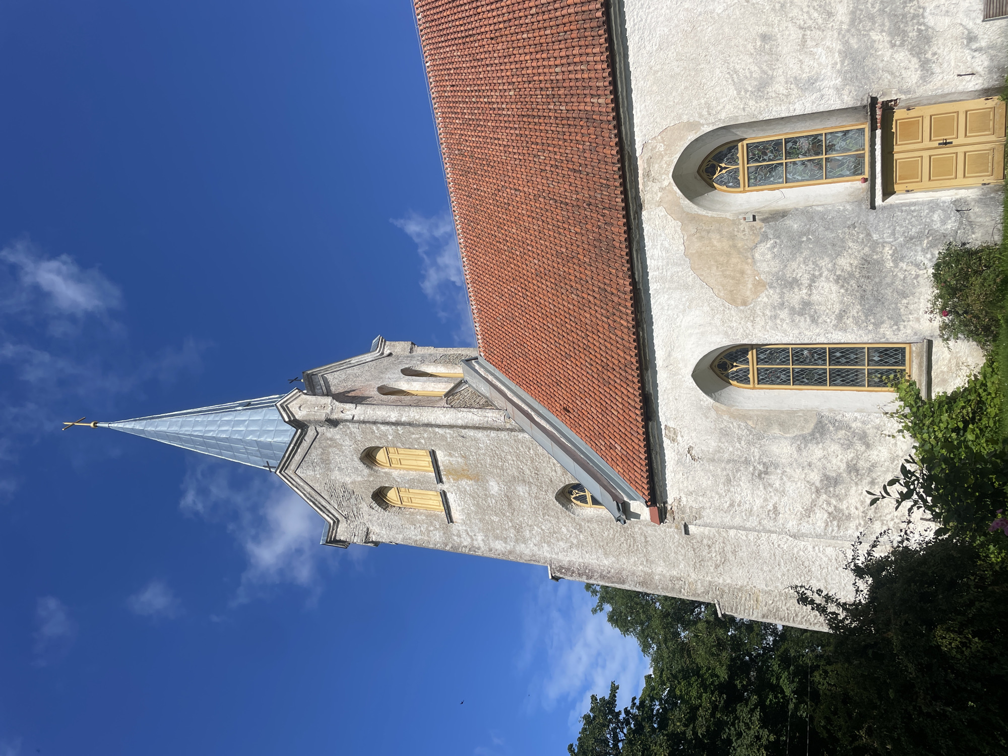 The image shows the Jõelähtme St. Nicholas' Church in Jõelähtme, Estonia, on a sunny day. The church is constructed of stone and has a prominent steeple topped with a golden cross. The roof is covered in red tiles, and several arched windows with yellow frames are visible on the building. Green trees and foliage surround the church. The sky is blue with scattered white clouds. No people are present in the image.