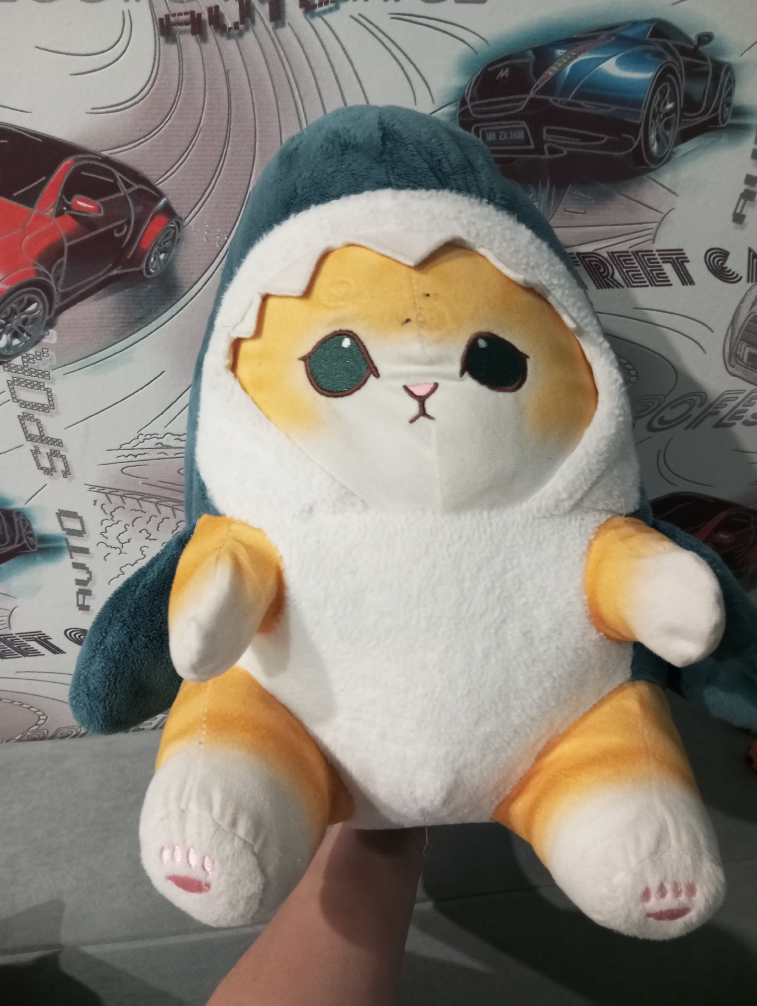The image shows a plush toy cat wearing a shark costume. The cat is white with orange markings on its paws and tail. It has large green eyes and a small pink nose and mouth. The shark costume is blue and white, with fins on the sides. The toy is sitting on a gray surface, possibly a couch or bed. In the background, there is a wallpaper with images of cars and street racing motifs. The image is taken from a slightly high angle, with a hand visible at the bottom, holding the toy. The overall lighting is soft, and the focus is on the plush toy. The specific location of Verkhnyaya Pyshma, Russia is not visually discernible from the image.