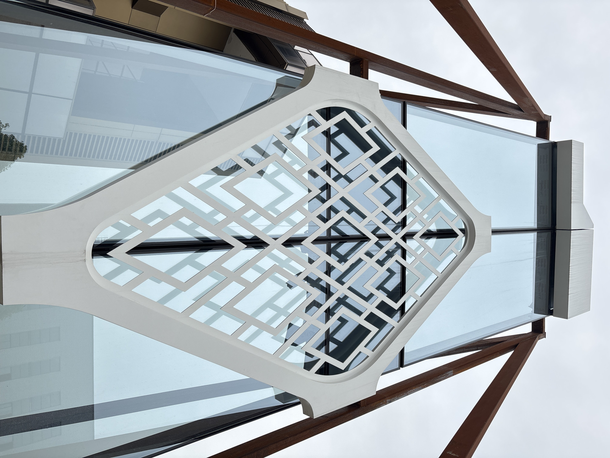 The image depicts a detail of a building facade in Chengdu, China, likely taken from a low angle looking upwards. The building features a geometric, latticework design in white, overlaid on large glass window panes. The lattice work is framed by a decorative border with curved edges. Visible in the reflection on the glass are other buildings, suggesting an urban setting. The building also has visible rusted-brown structural supports or beams. The sky in the background is overcast and grey. The scene is an architectural detail showing a modern design with traditional elements.