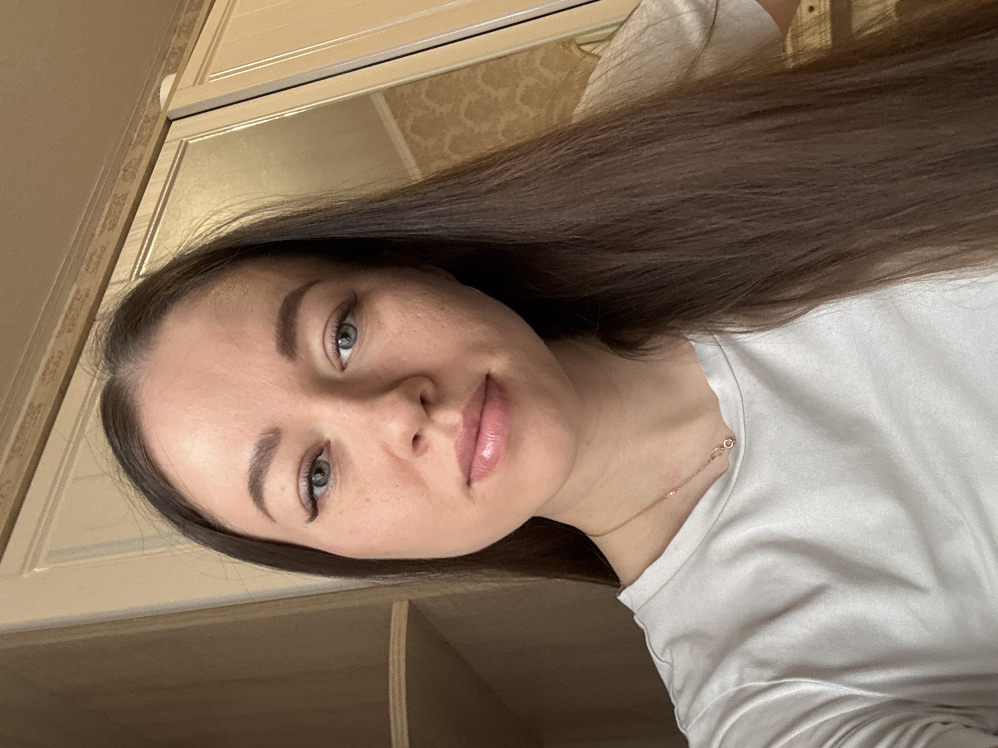 A woman with long brown hair, green eyes, and fair skin is shown in a close-up, head-and-shoulders shot. She is wearing a white t-shirt and a delicate gold necklace. The background includes cabinet doors with decorative detailing. The location is Ферма, Russia.