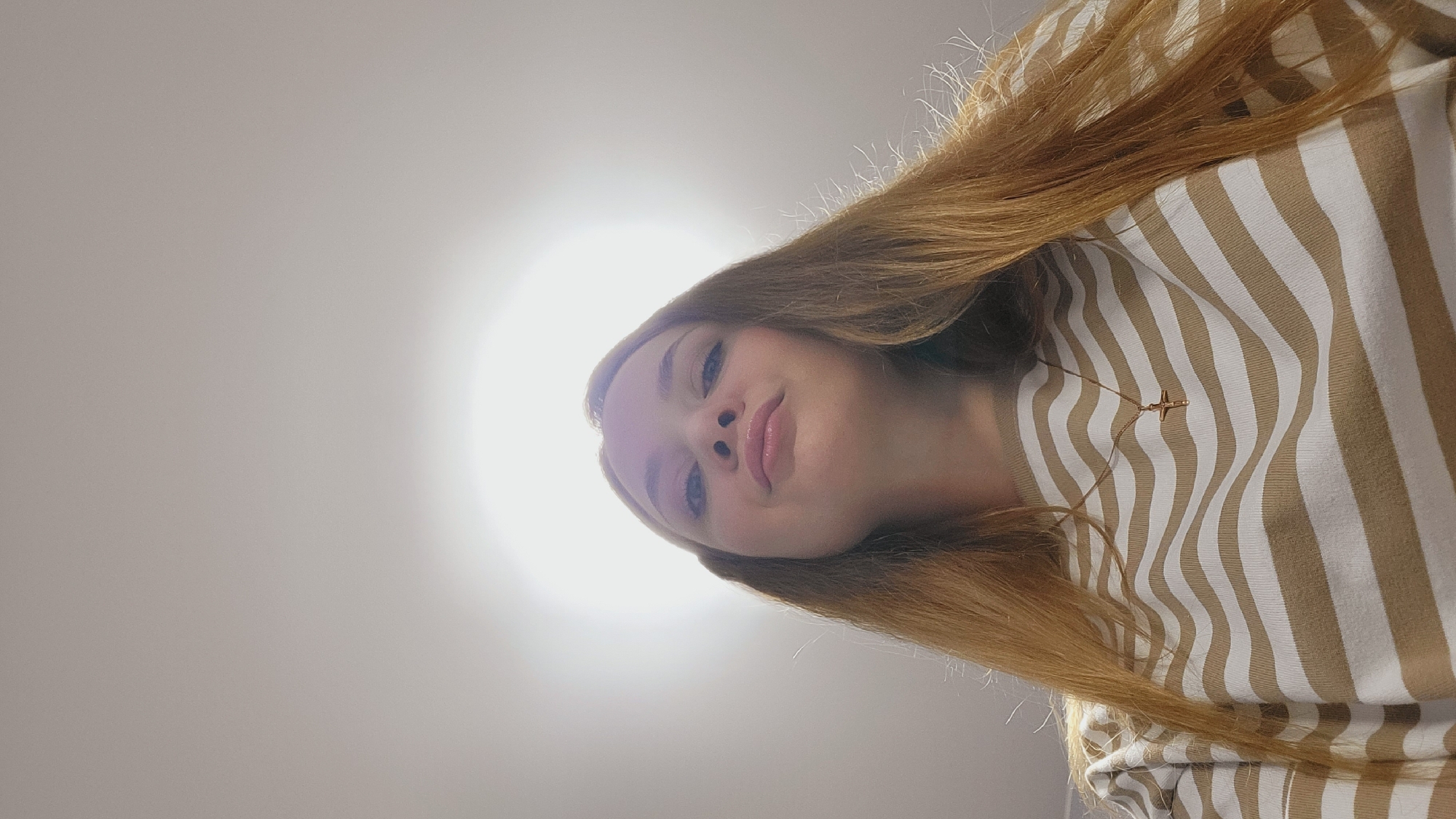 A woman with long, reddish-blonde hair is positioned at an angle, looking down into the camera. She has fair skin, blue eyes, and glossy lips. She is wearing a tan and white striped long-sleeved shirt. A small gold-colored pendant hangs from a delicate chain around her neck. The background is a bright, uniform light, possibly from a ceiling light. The image perspective suggests the photograph was taken from above. There is no indication of the location being Chaikovsky, Russia in the image.