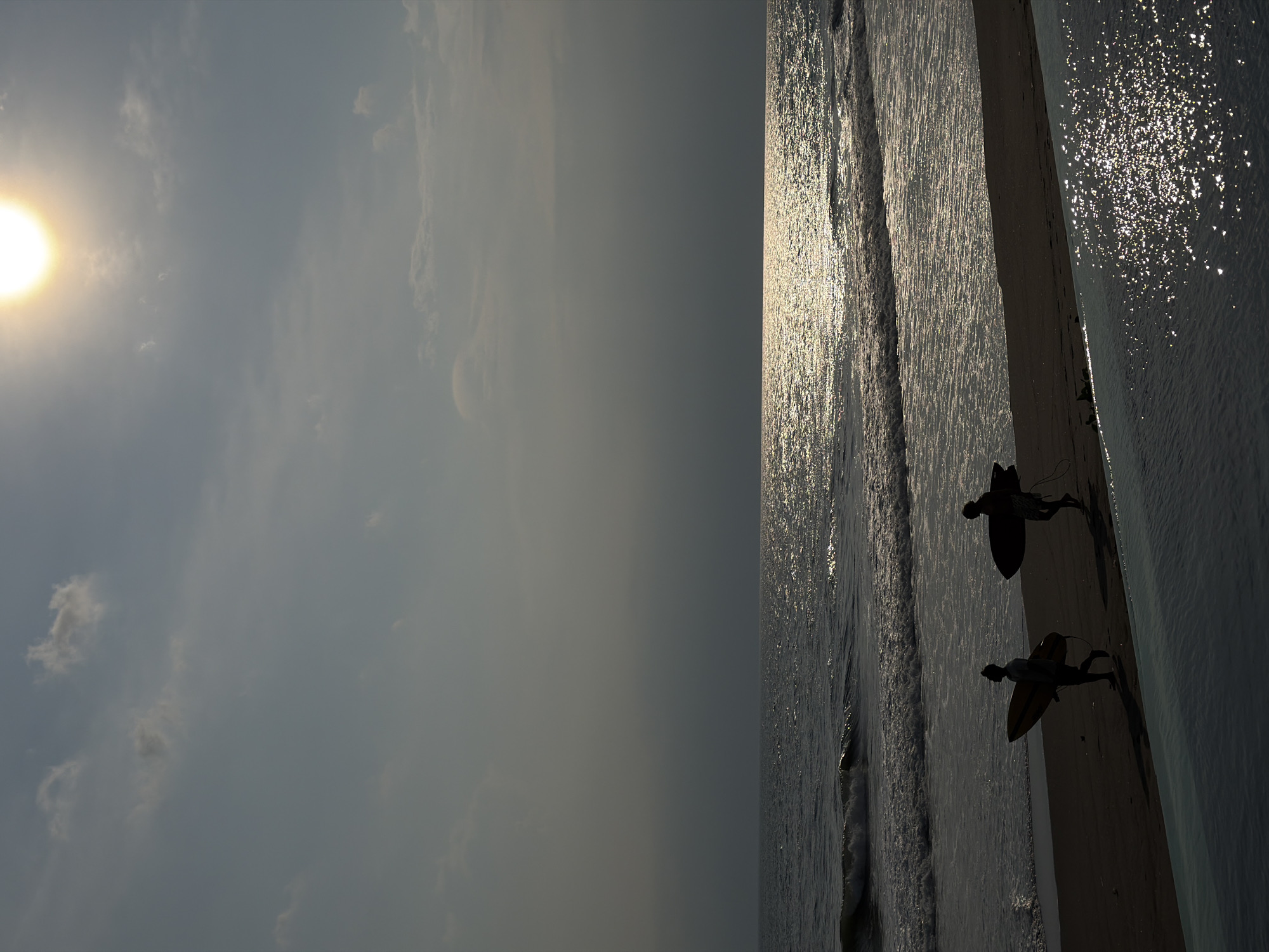 A backlit coastal scene is depicted in Belalang, Indonesia. The bright, intense sun, positioned in the upper right, illuminates a hazy, pale blue-grey sky with scattered clouds. The ocean to the right exhibits a broad, sparkling path of sunlight reflecting on its surface, extending to the horizon. Small waves break along the dark sand shoreline. Two individuals, appearing as dark silhouettes, traverse the beach from left to right, each carrying a surfboard. The surfboard carried by the person on the left is visibly lighter in tone than the dark board held by the person on the right. In the immediate foreground, a body of water, likely an infinity pool, glistens with numerous bright reflections from the overhead sun.