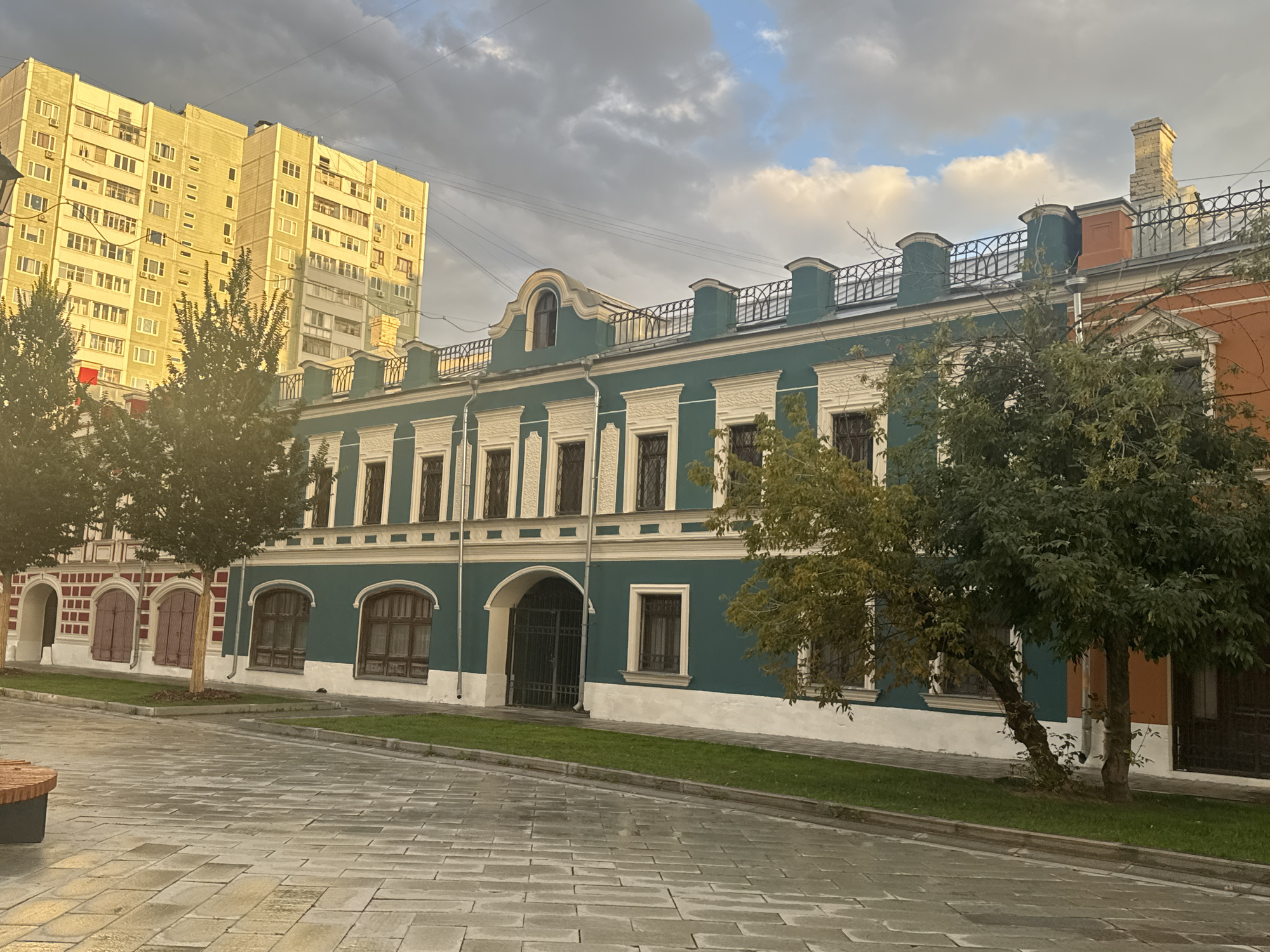 The image depicts a street scene in Moscow, Russia, bathed in the soft light of what appears to be late afternoon or early evening, with a partly cloudy sky. Dominating the right side of the frame is a low-rise building with a distinctive teal facade, adorned with white decorative elements around the windows and along the roofline. This building has a series of arched windows and a large arched entryway with a black metal gate. To the left of this teal building, and slightly set back, is a section of an older building with red brickwork and burgundy shutters. Further to the left, a modern, tall, pale yellow apartment building rises prominently against the sky. In the foreground, a paved walkway with a stone-like pattern stretches across the scene. Green grass borders the sidewalk in front of the buildings. Two mature trees with full green foliage are positioned along the sidewalk, partially obscuring the teal building. The overall impression is one of a mix of architectural styles within a city environment.