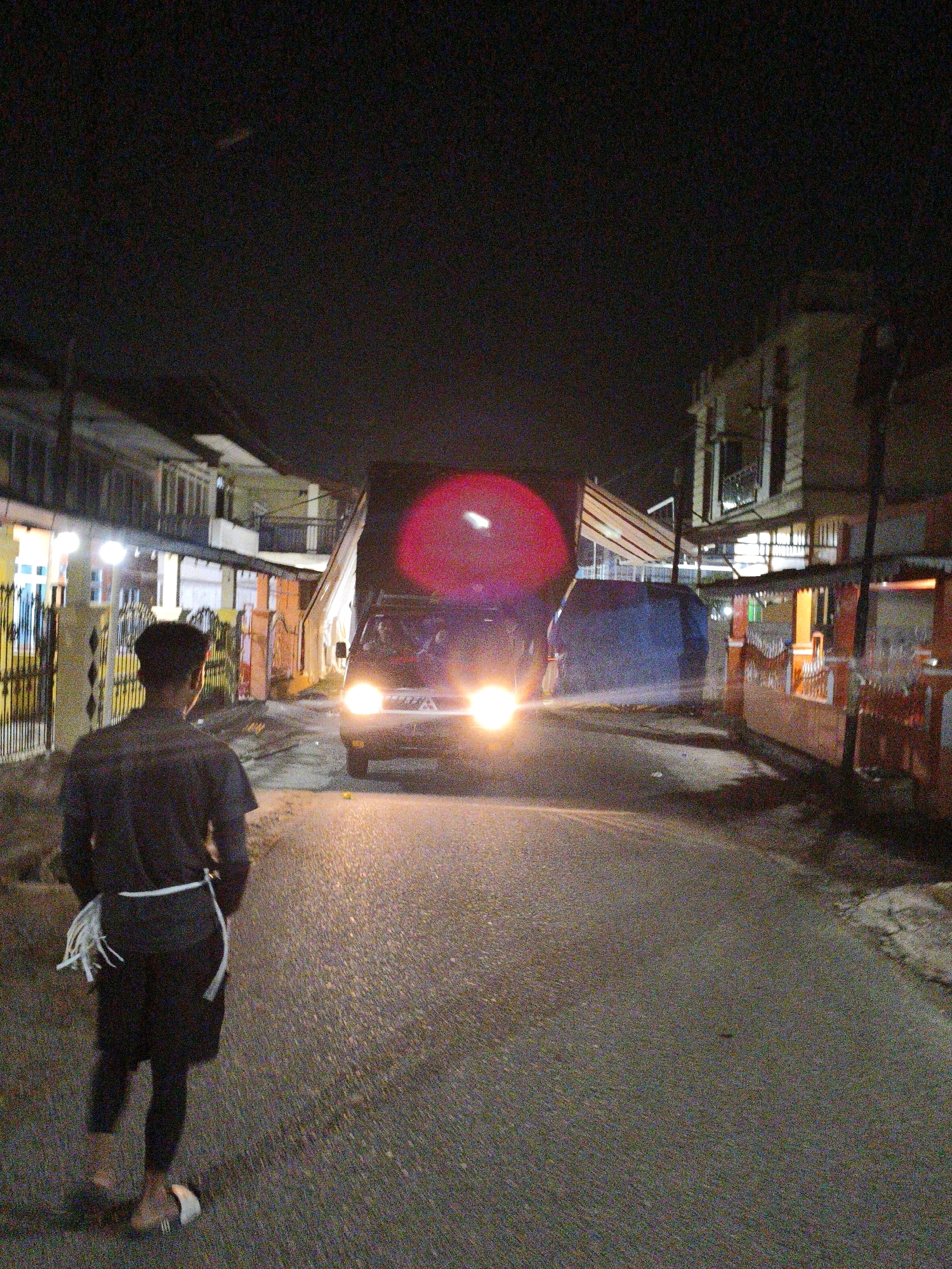 A lone individual, seen from behind, walks away from the camera down a dimly lit street in Koto Baru Semurup, Indonesia. The person wears a dark t-shirt, dark shorts over dark leggings, and sandals. A white sash or rope is tied around their waist, with loose ends hanging. The street is paved and appears wet, reflecting the headlights of an approaching truck. The truck, with its bright headlights on, is a dark color and has a large red circular object or light reflection on its front. On either side of the street are buildings with lights on. The overall scene is nocturnal, with visible grain in the photograph.