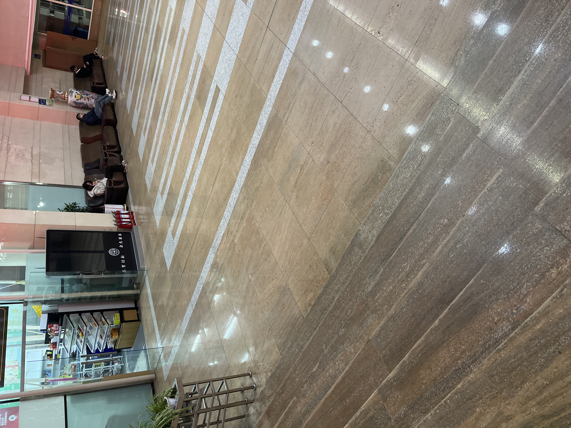 The image depicts an interior space in Changchun City, China, likely a waiting area within a transportation hub or a commercial building. The floor is a combination of light brown and grey stone tiles, with white, rectangular inlays running parallel in the light brown section. Seating is available with a row of brown, cushioned chairs positioned against a wall. Several individuals are visible: some are sitting and appear to be waiting, others are reclining on the chairs, potentially resting. A small convenience kiosk, enclosed by glass, displays various retail goods. There are two fire extinguishers, in front of the kiosk. A metal railing is located towards the bottom of the image.